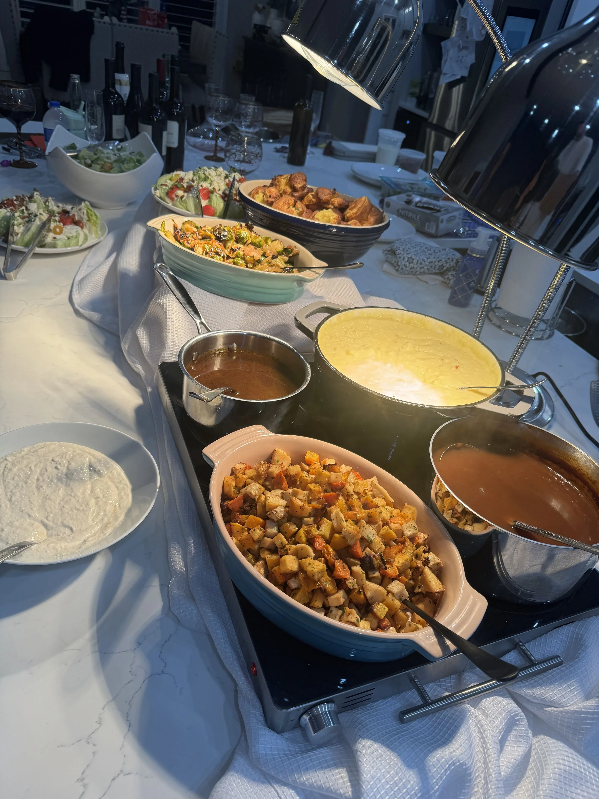 A buffet table with various dishes including roasted potatoes, salads, and casseroles. There are industrial-style lamps overhead, and the background features a dining area with wine bottles and glasses.