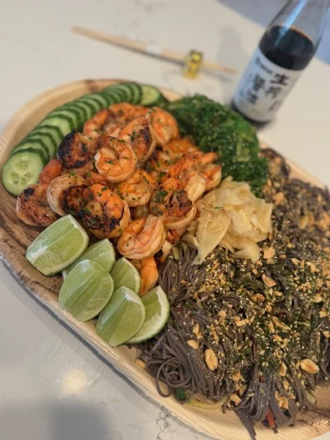 A wooden platter holding grilled shrimp, sliced cucumbers, lime wedges, broccoli, pickled ginger, black noodles topped with crushed nuts and vegetables, and a soy sauce bottle in the background.