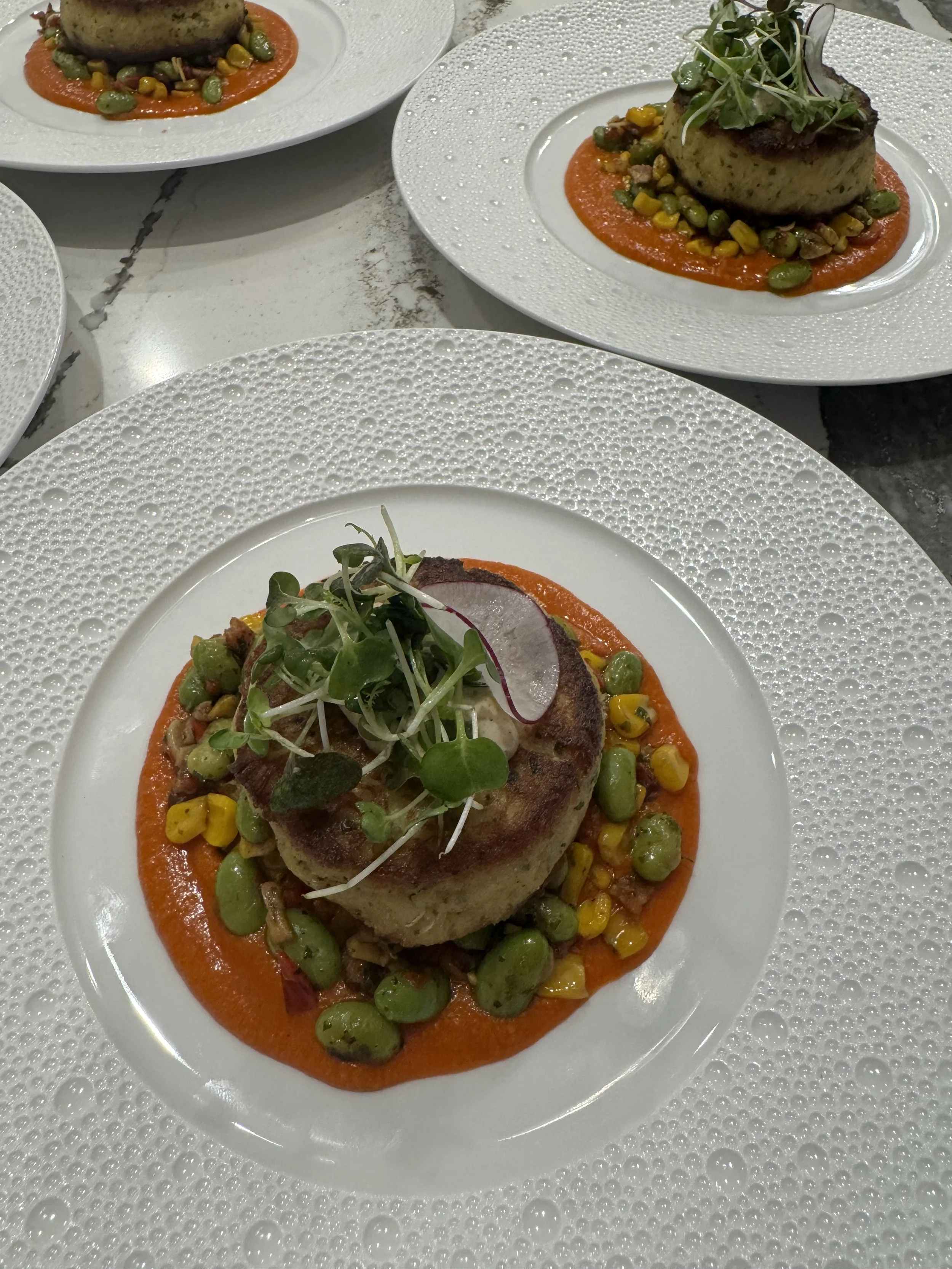 Plate with a baked potato topped with microgreens, served on a bed of colorful corn and green peas on an orange sauce, all on a white textured plate.