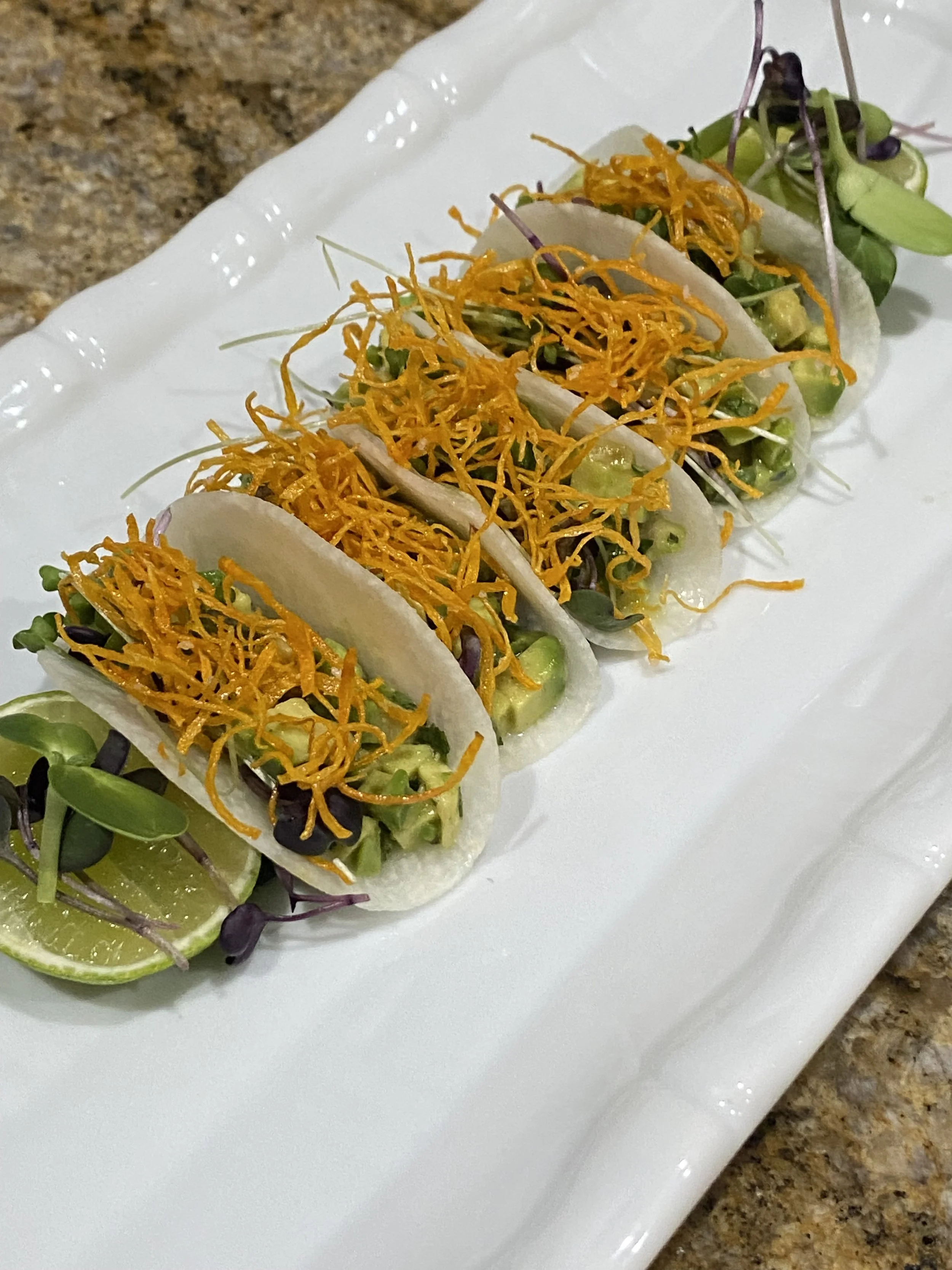 Three soft shell tacos filled with microgreens, avocado, and shredded cheese, served on a white plate with lime wedges.