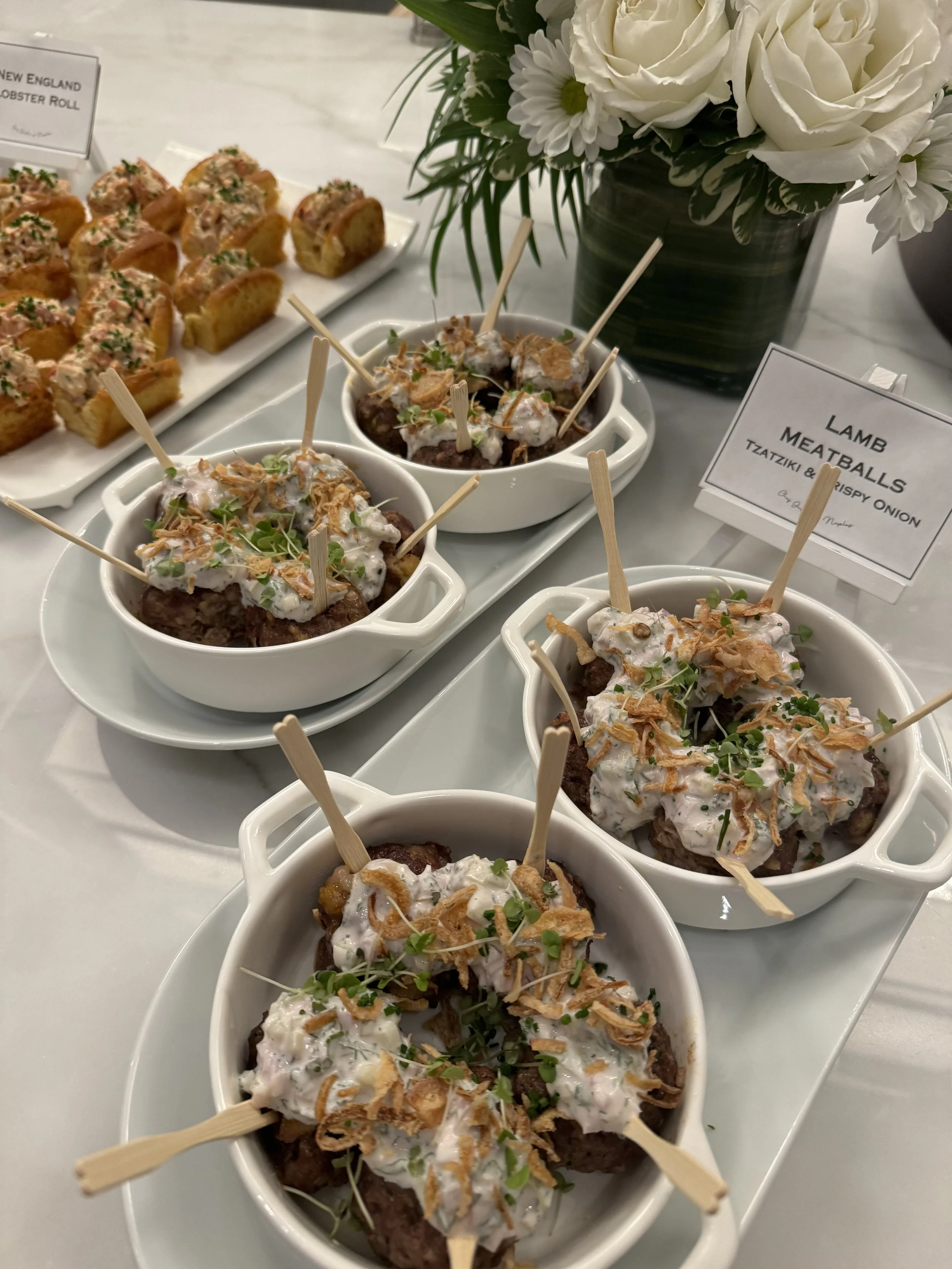 Small white bowls filled with lamb meatballs topped with a creamy sauce, crispy onions, and green herbs, arranged on a white tray. A sign indicates the dish is lamb meatballs with tzatziki and crispy onion. In the background, a large bouquet of white