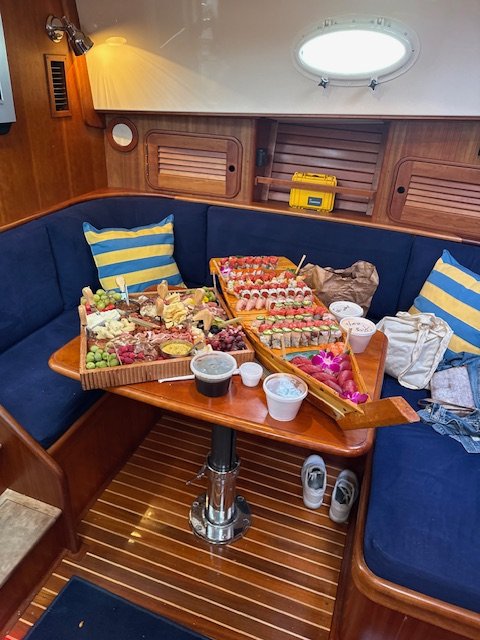 Boat interior with a curved navy blue cushioned seating area, a wooden table with assorted snacks and drinks, striped pillows, and wood-paneled walls.