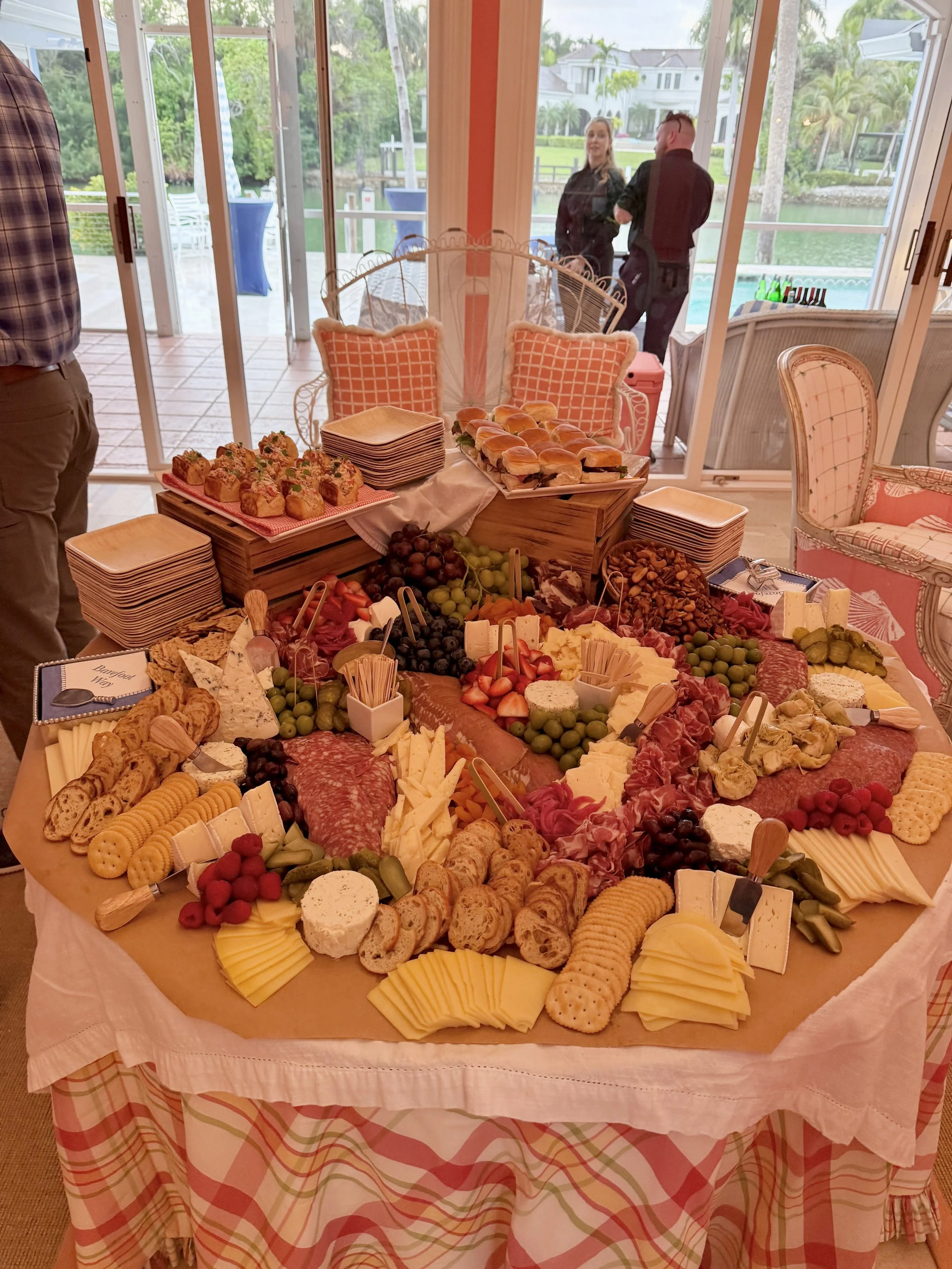A cheese and charcuterie platter with assorted cheeses, crackers, grapes, strawberries, and meats, arranged on a round table with a pink and white plaid tablecloth. In the background, there are decorative chairs and a view of a lake or river outside 