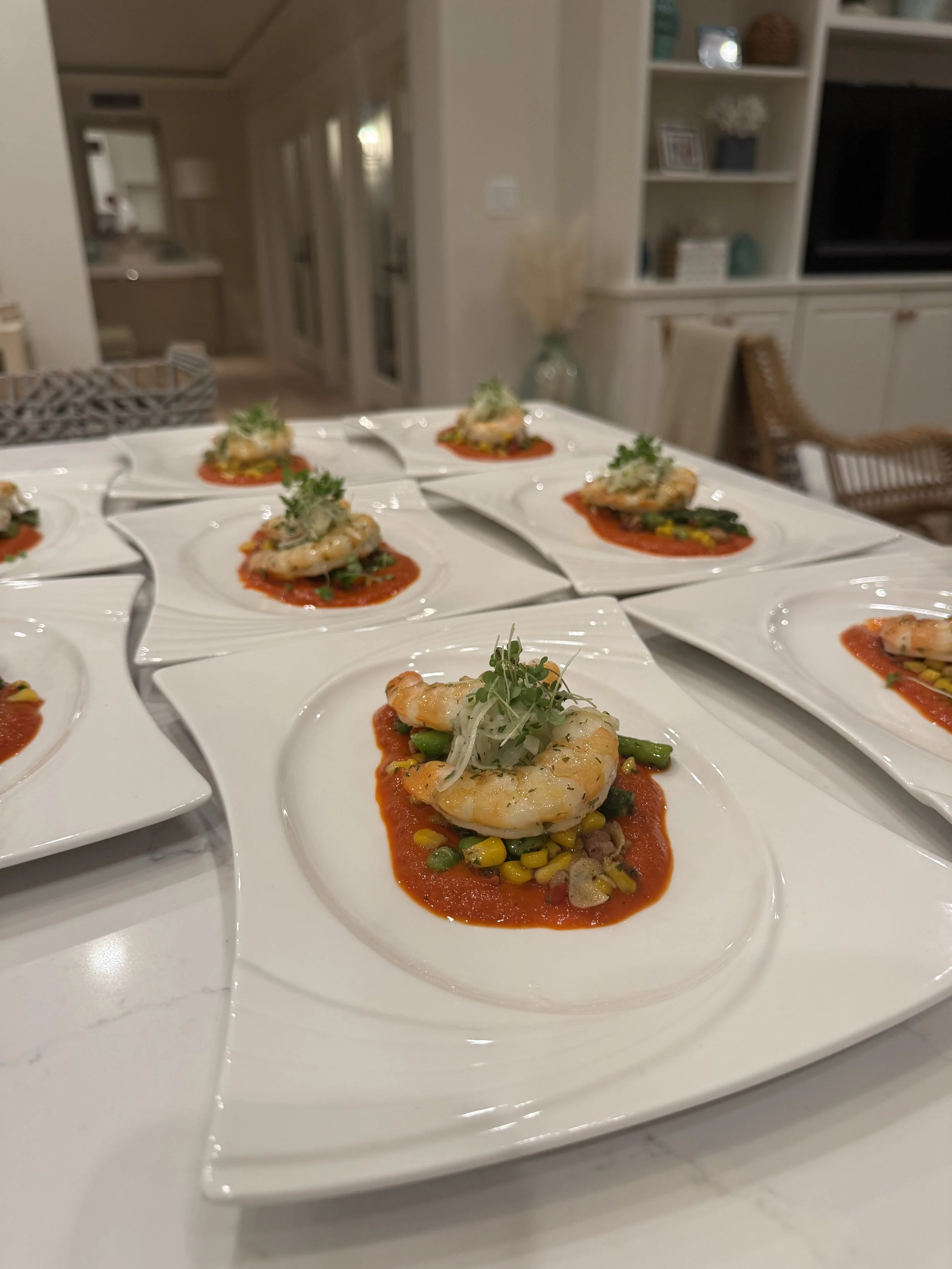 Multiple white square plates with shrimp on a bed of tomato sauce, topped with microgreens, with a background of a modern kitchen and dining area.