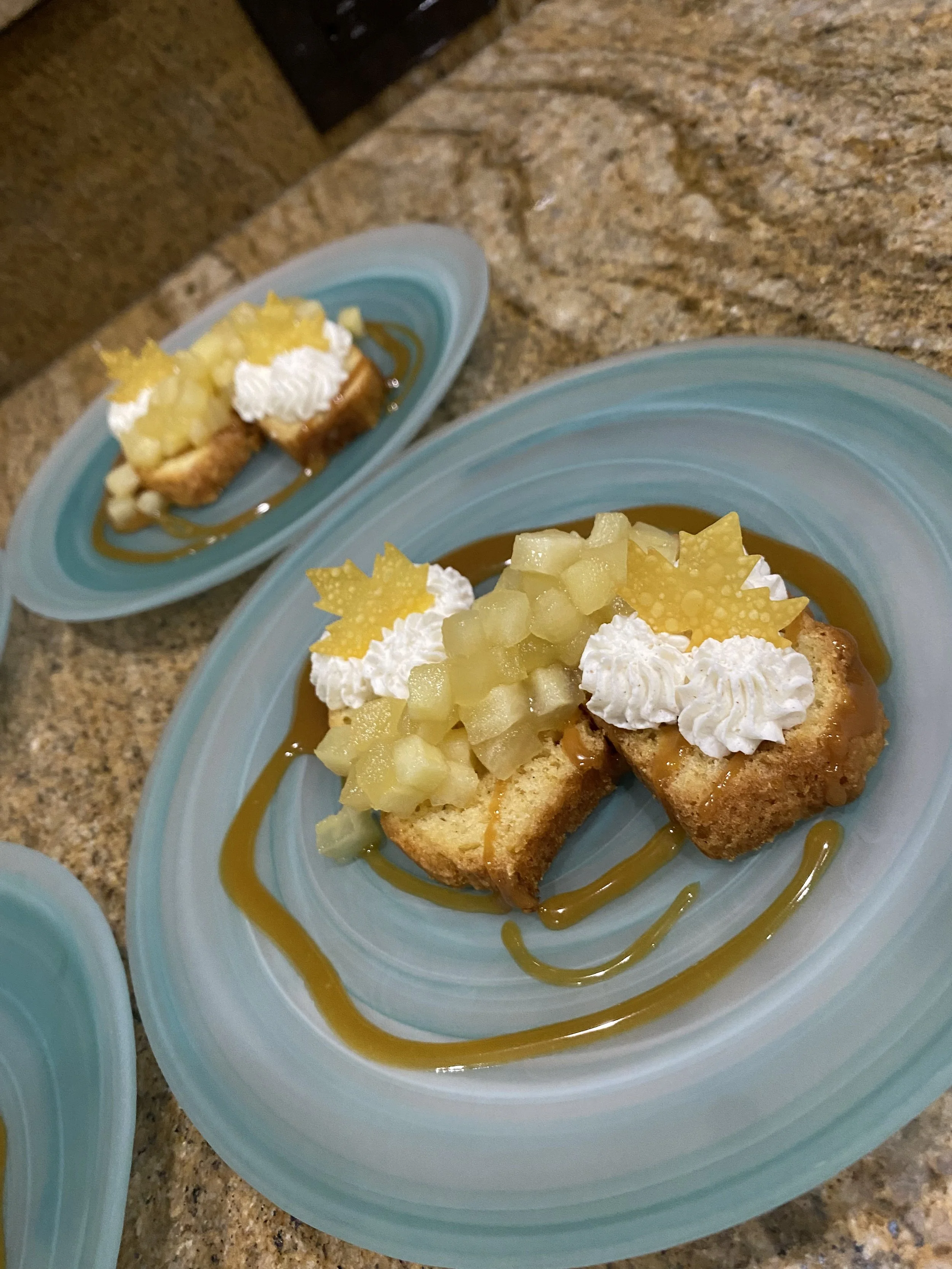 Close-up of a dessert on a blue plate, featuring a slice of cake topped with whipped cream, diced yellow fruit, and decorative yellow wafer stars, with caramel drizzle and a caramel background.