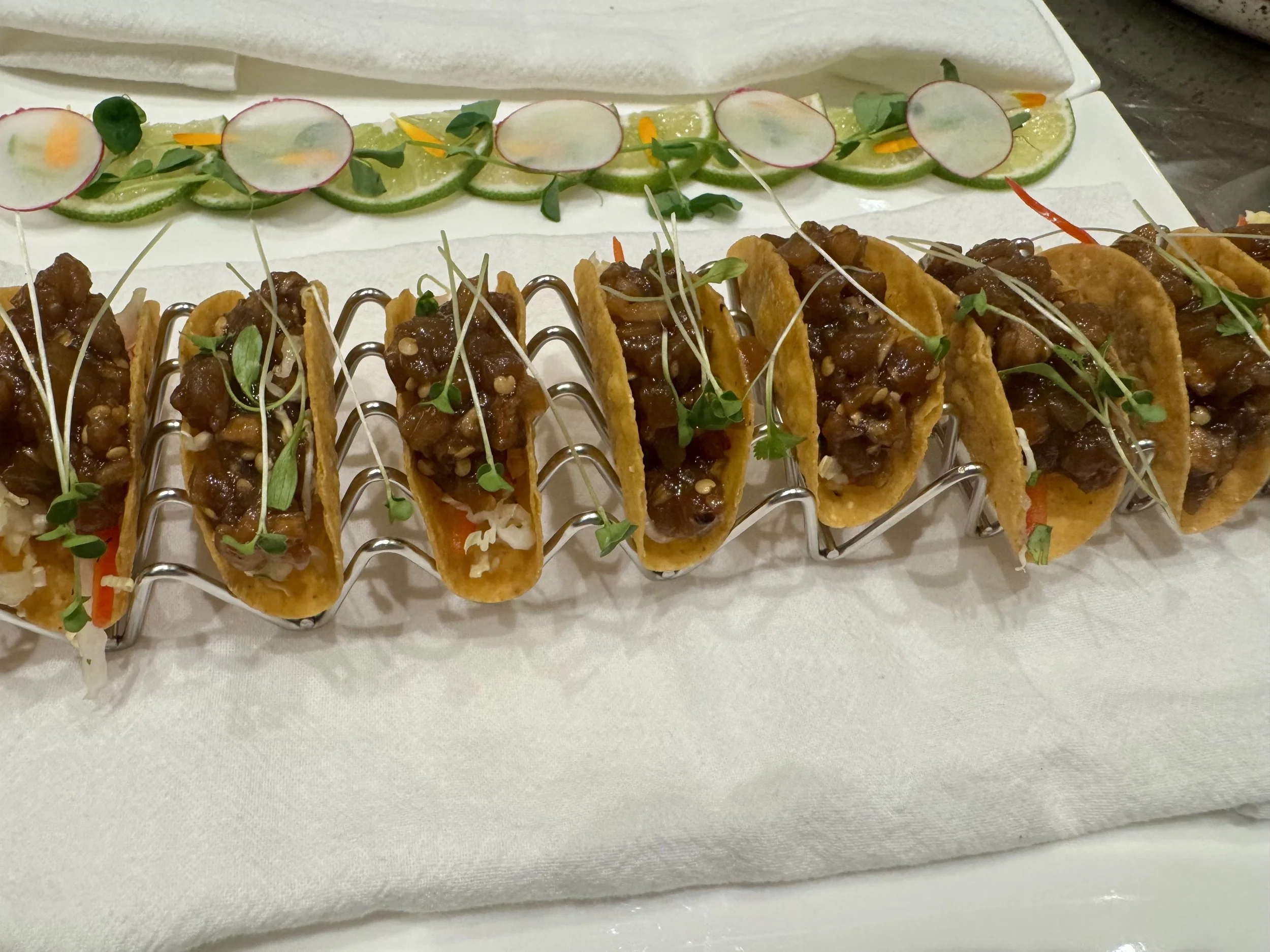 Close-up of a row of mini tacos filled with meat, topped with microgreens, on a metallic rack.