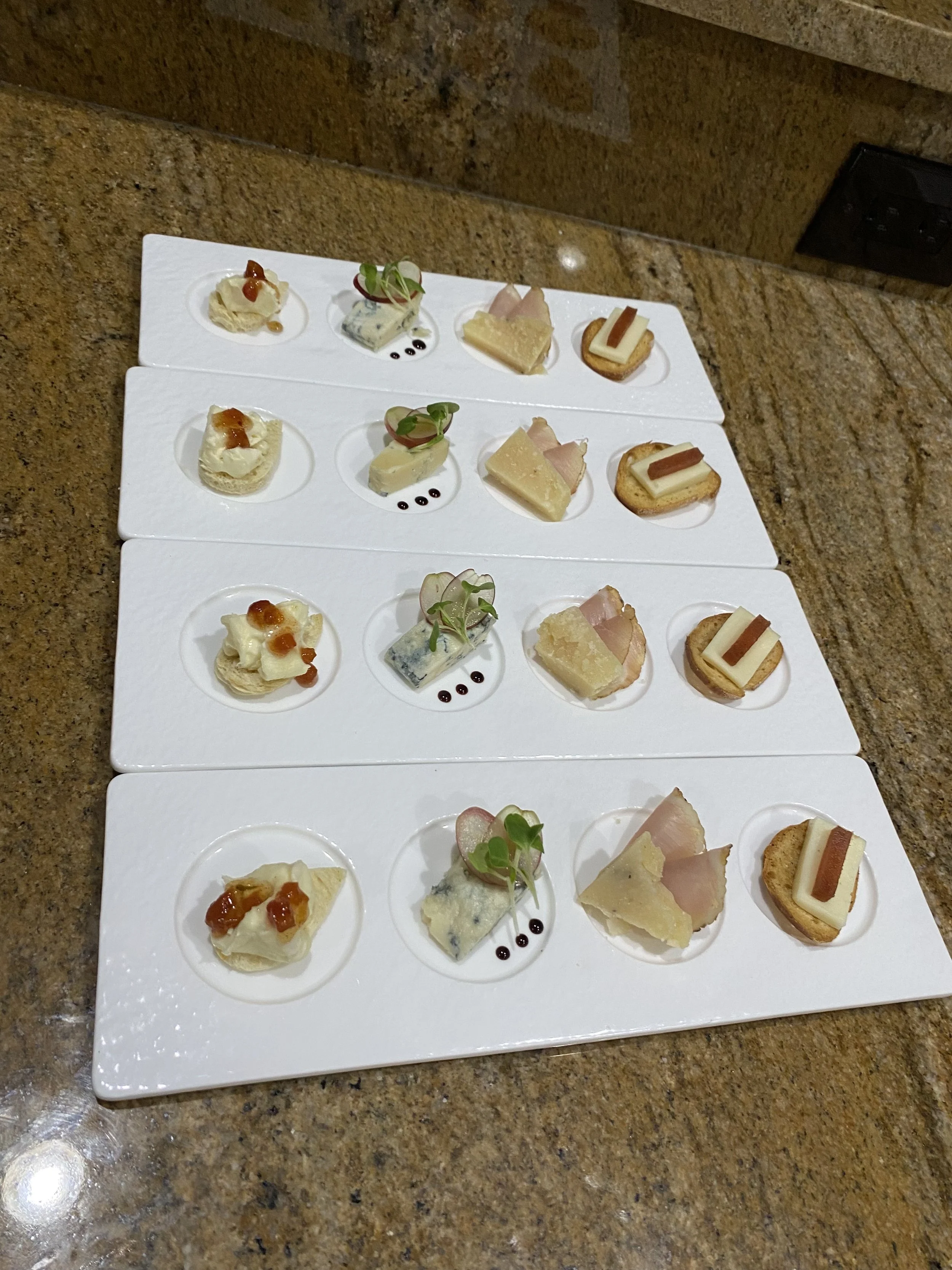 A white tray with three rows of four small canapé snacks, including cheese, meats, and garnishes, placed on a brown granite countertop.