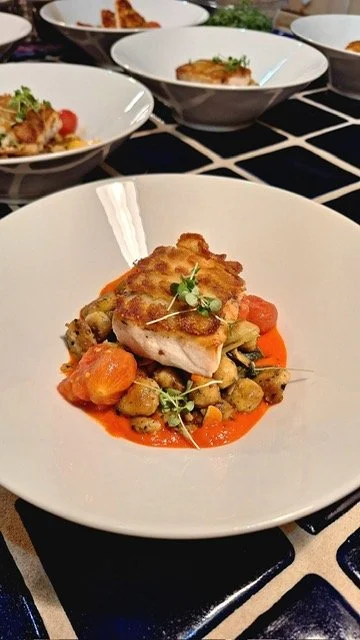 A plated dish with a piece of cooked fish on a bed of roasted vegetables with a red sauce, garnished with microgreens.