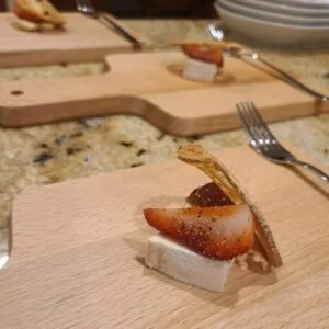 Close-up of a dessert with a strawberry slice, a rectangular piece of cheesecake, and a cookie on a wooden serving board, with a fork nearby.