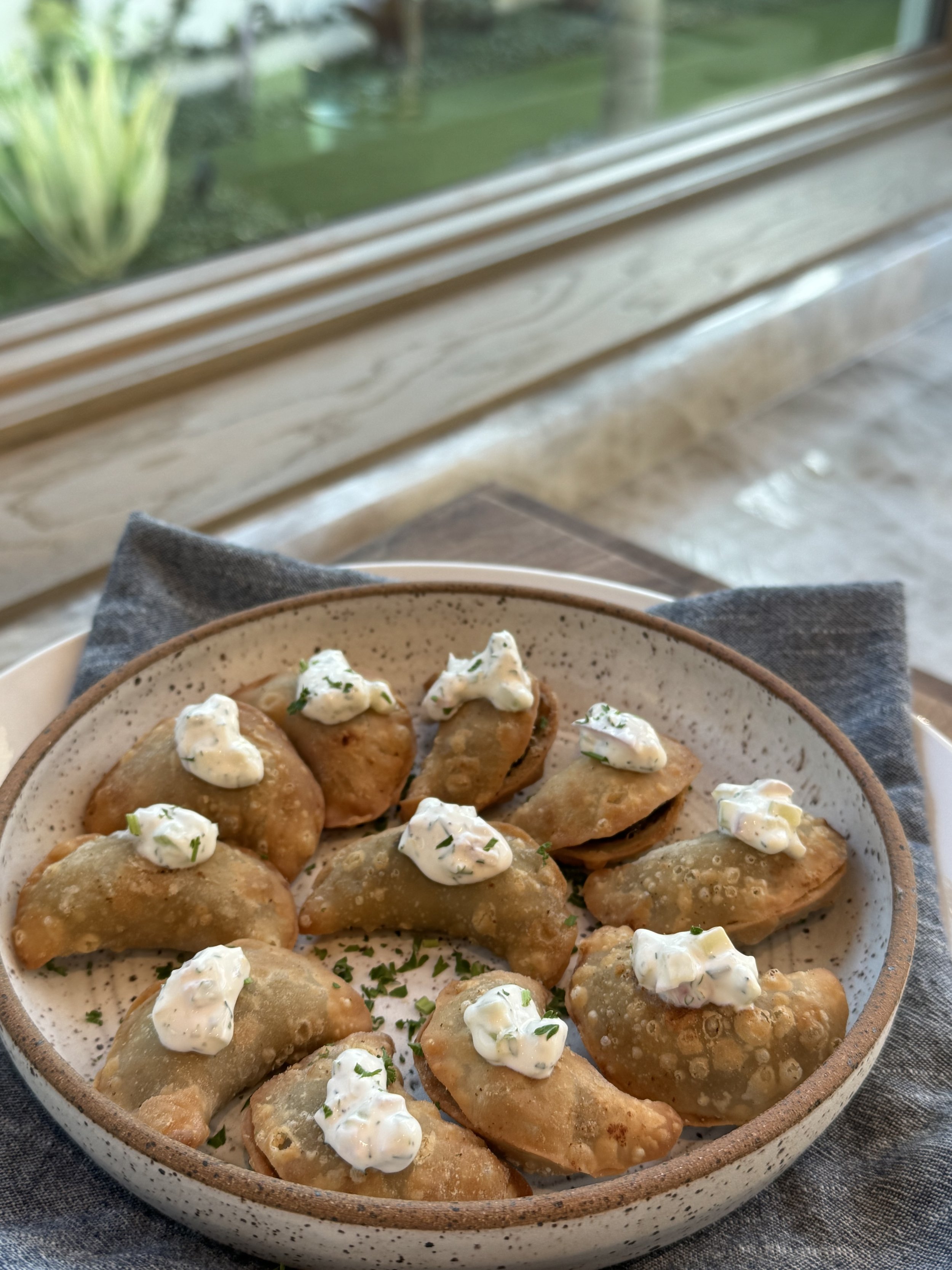 A bowl of potato skins topped with sour cream and chopped herbs, placed on a dark cloth near a window with a garden view.