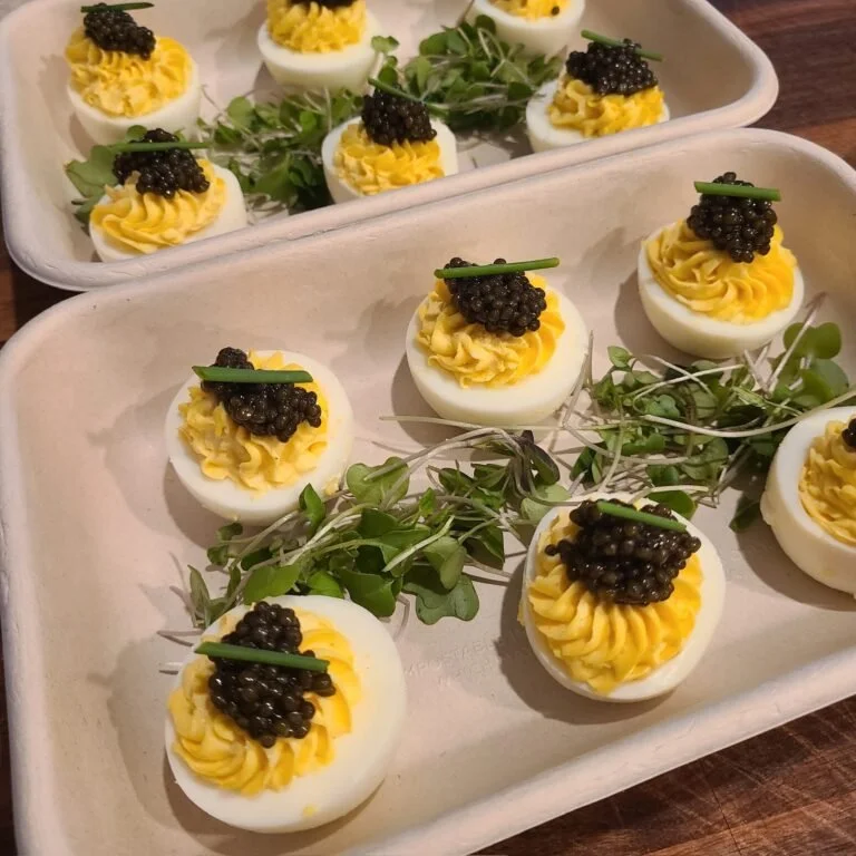 Deviled eggs garnished with black caviar and chives, arranged on a white tray with greenery as decoration.
