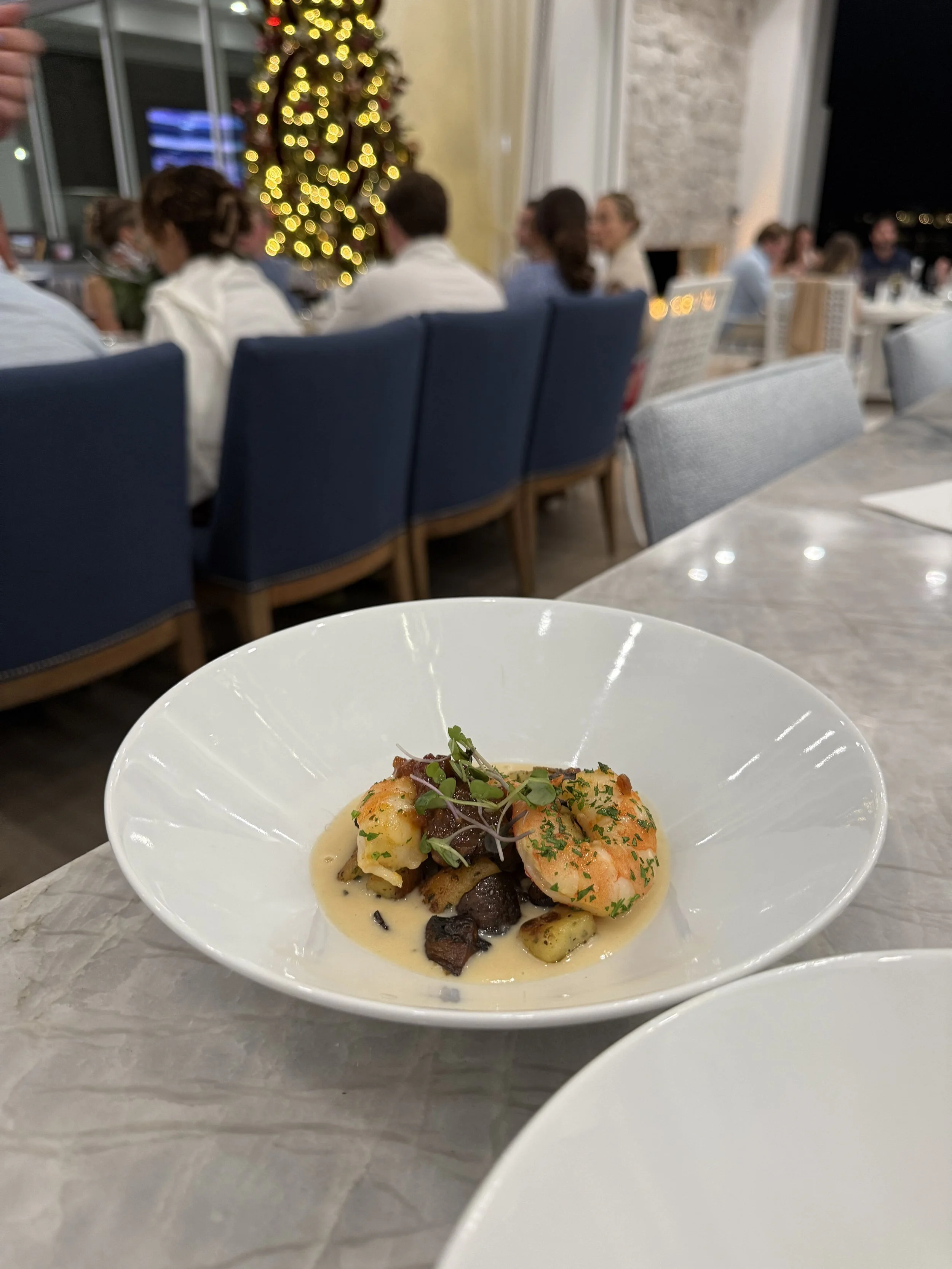 A white bowl of seafood with shrimp, garnished with herbs and microgreens, on a marble table, with a blurred dining room and a decorated Christmas tree in the background.