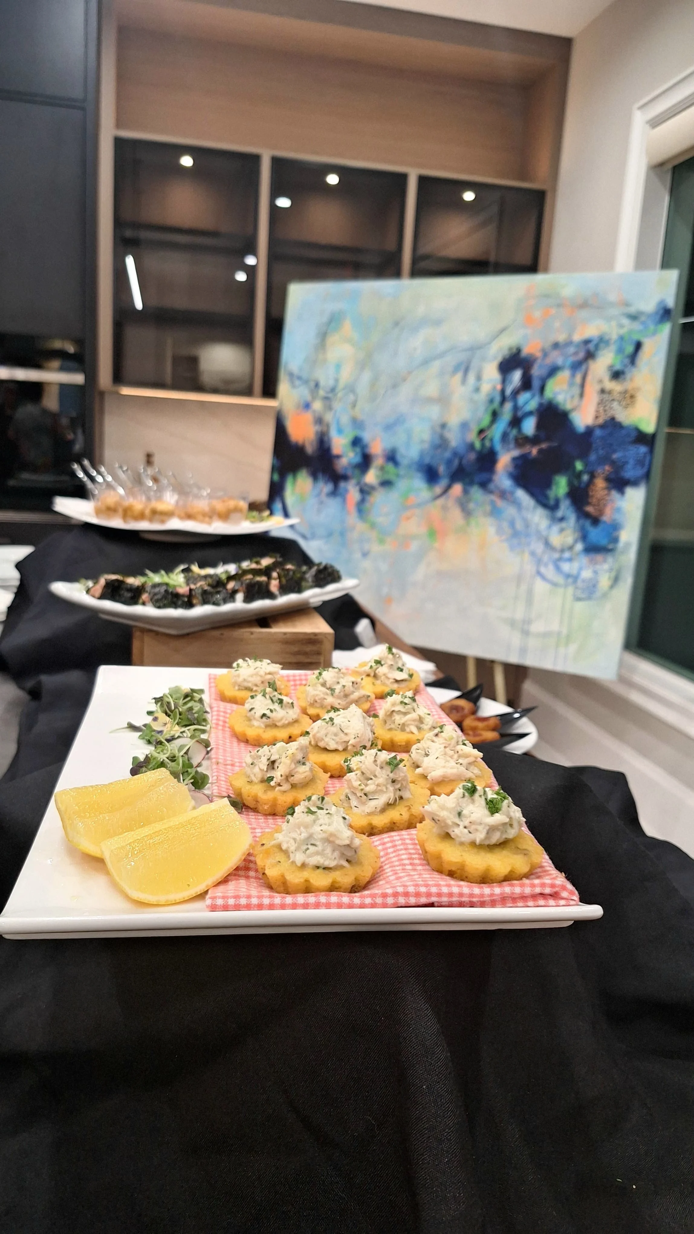 A food buffet display in a modern room, featuring a white platter with lemon wedges and square-shaped bites topped with creamy mixture, a second black platter with sushi rolls, and a third platter with assorted appetizers, all arranged on a black tab