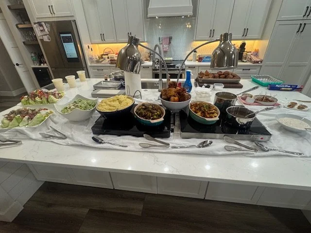 Kitchen island with various dishes and beverages, including salads, mashed potatoes, fried chicken, and pies, with heat lamps overhead.