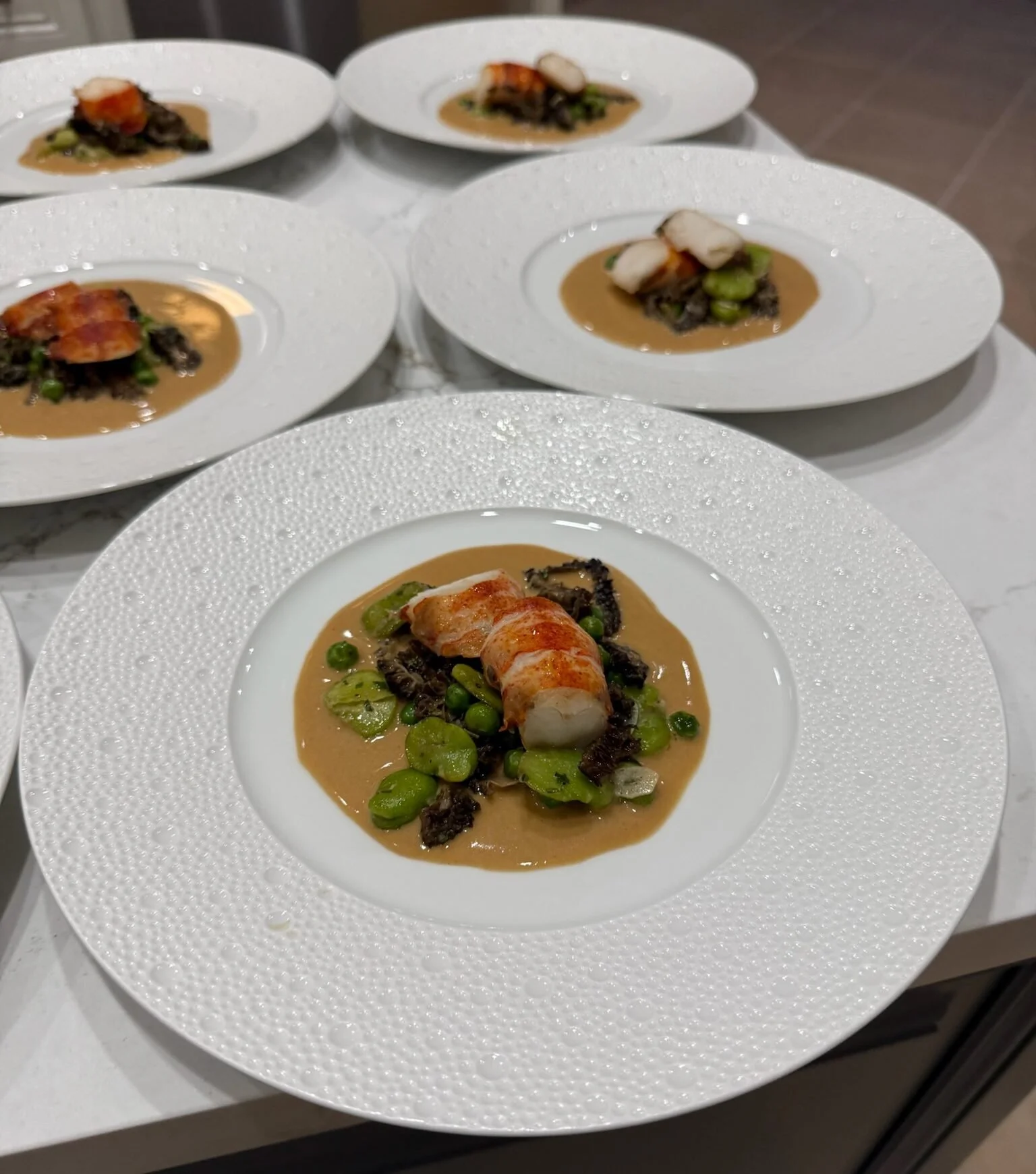 Five plates of gourmet food with a creamy sauce, vegetables, and lobster tail on white textured plates.