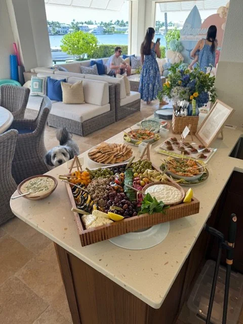 A buffet table with a variety of snacks, including grapes, sliced bread, crackers, and dips, set up in a bright, modern living space with large windows, couches, and people in the background.