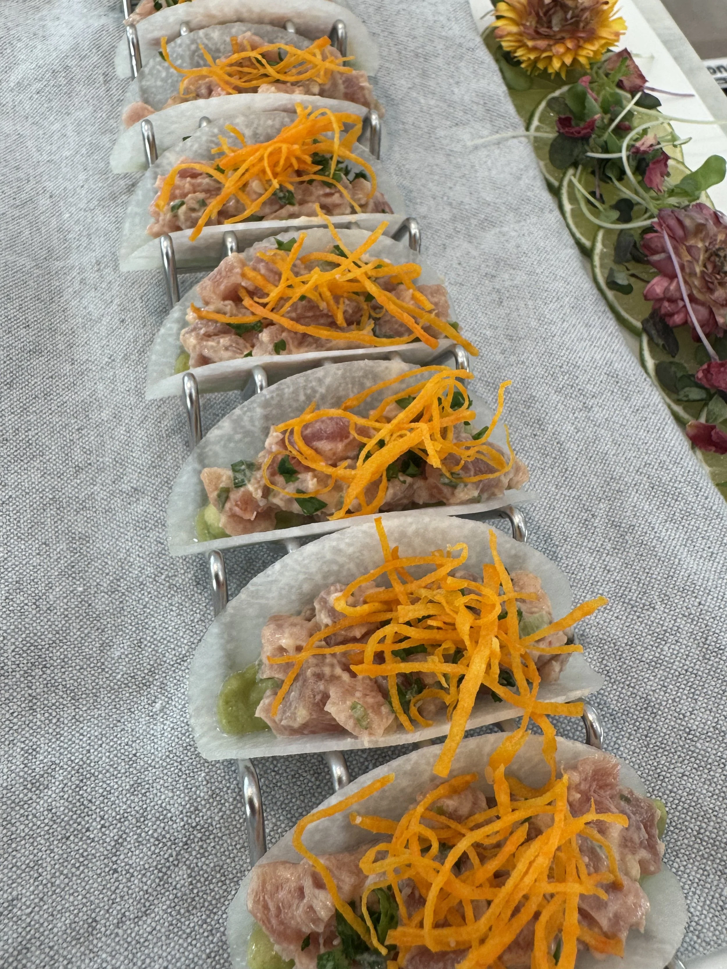 Close-up of a line of spring rolls topped with shredded orange carrots, placed on a paper-lined tray, with a floral garnish and lemon slices in the background.