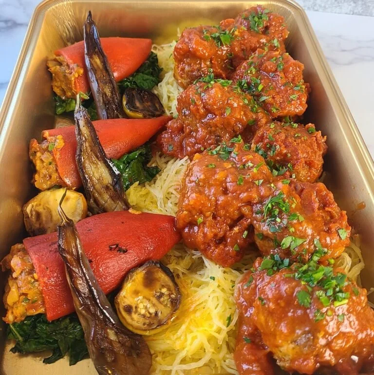 A dish with breaded meatballs covered in tomato sauce garnished with chopped parsley, served over shredded cheese, with roasted eggplants, red bell peppers, and sautéed greens.