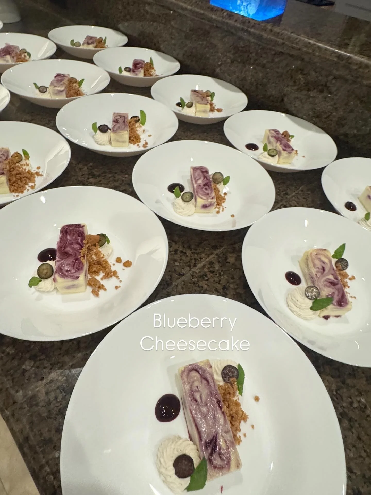 Multiple white plates with blueberry cheesecake, garnished with blueberries, mint leaves, small dollops of cream, and small dots of berry sauce, arranged on a dark granite countertop.
