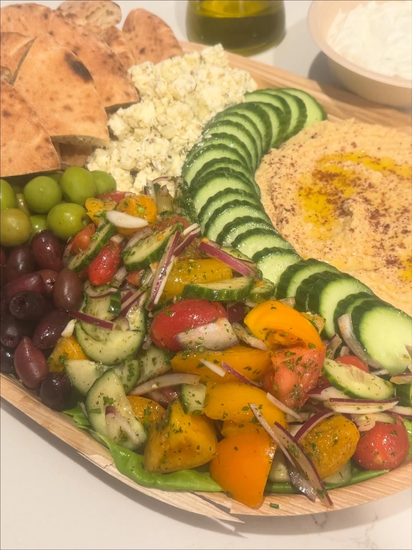 A wooden platter with assorted fresh vegetables including sliced cucumbers, cherry tomatoes, yellow peppers, red onions, green olives, and black olives, along with a side of crumbled cheese, pita bread, and a bowl of hummus, with a bottle of olive oi