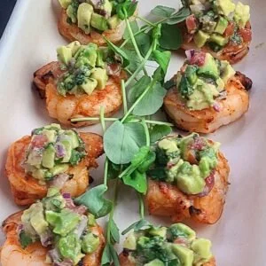 Six pieces of baked chicken topped with diced avocado and red onion, garnished with microgreens, arranged on a white platter.