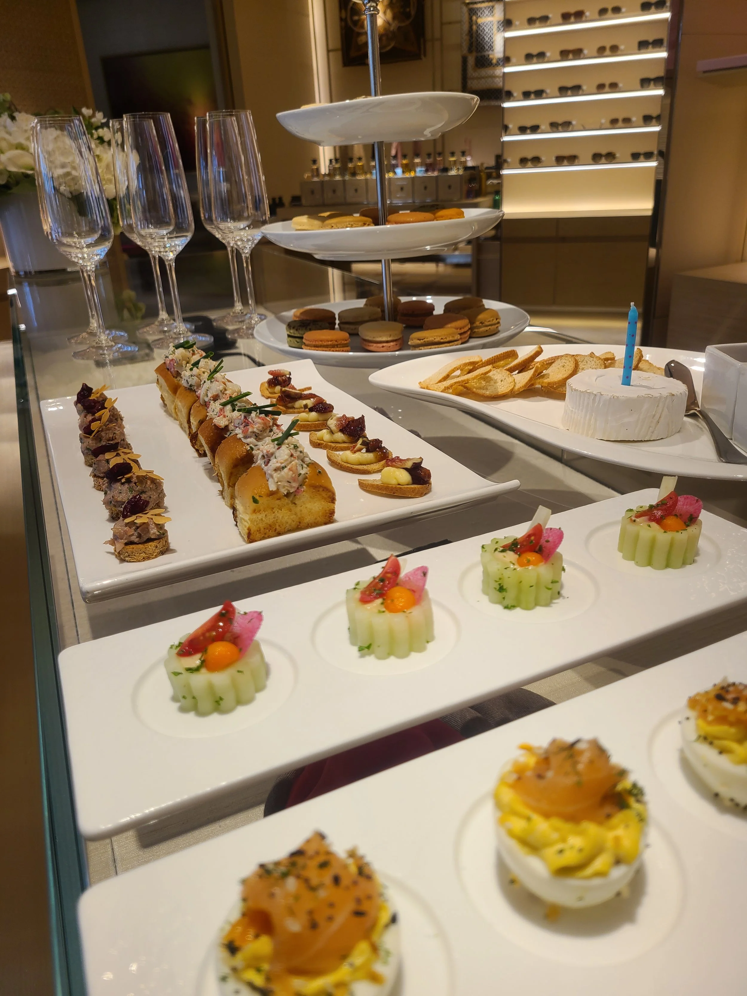 A display of assorted desserts and snacks, including small sandwiches topped with garnishes, colorful macarons on a tiered tray, and appetizer cups with vegetables and sauces, presented on white plates at a catering event.
