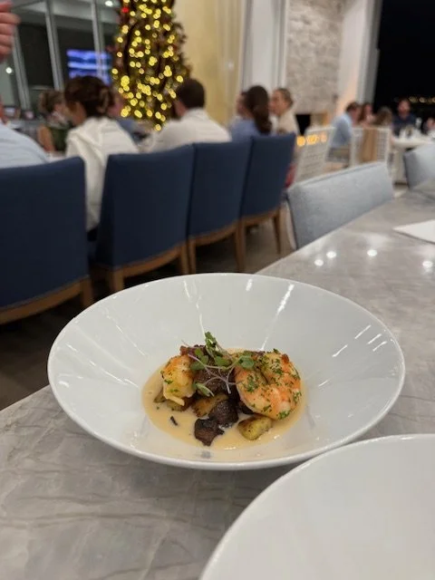A plate of gourmet seafood with shrimp, garnished with microgreens, on a marble table in a restaurant decorated with Christmas lights in the background.