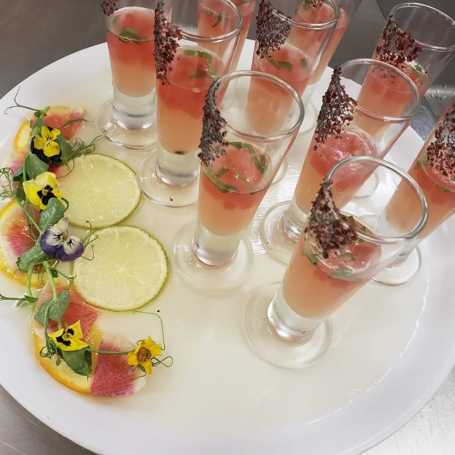A white platter holding multiple shot glasses filled with a pink and white layered beverage, garnished with greenery and sugar on the rim. The platter is decorated with slices of citrus fruits and edible flowers.