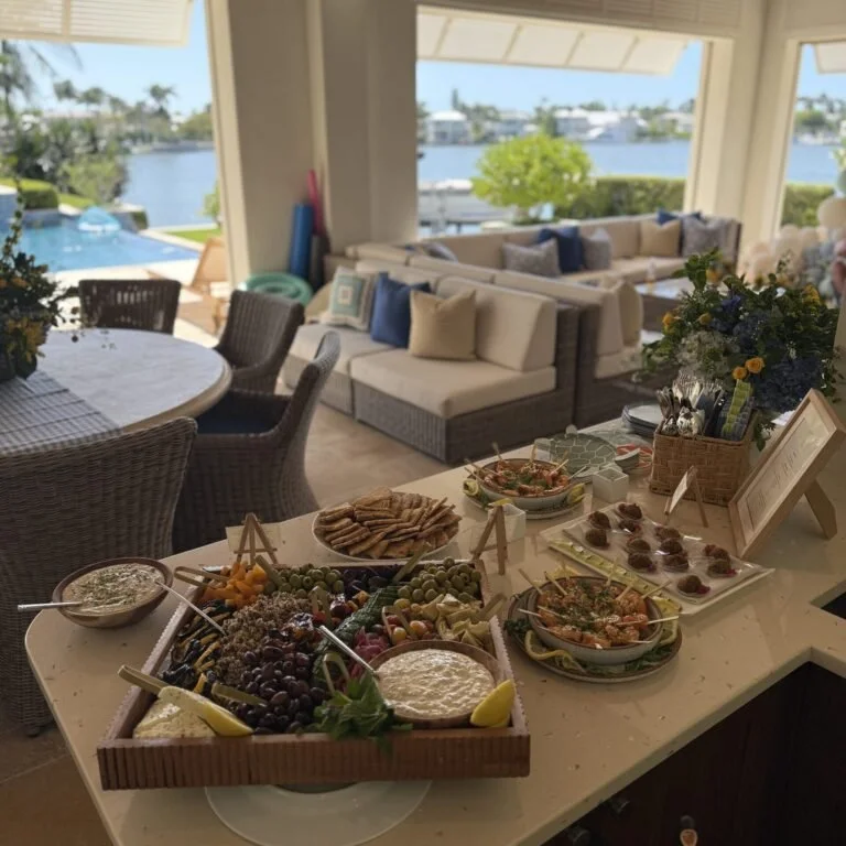 A buffet table in a bright, open living space overlooking water with a pool and boat dock. The table has assorted snacks including cheese, grapes, crackers, and dips, with a floral arrangement nearby.