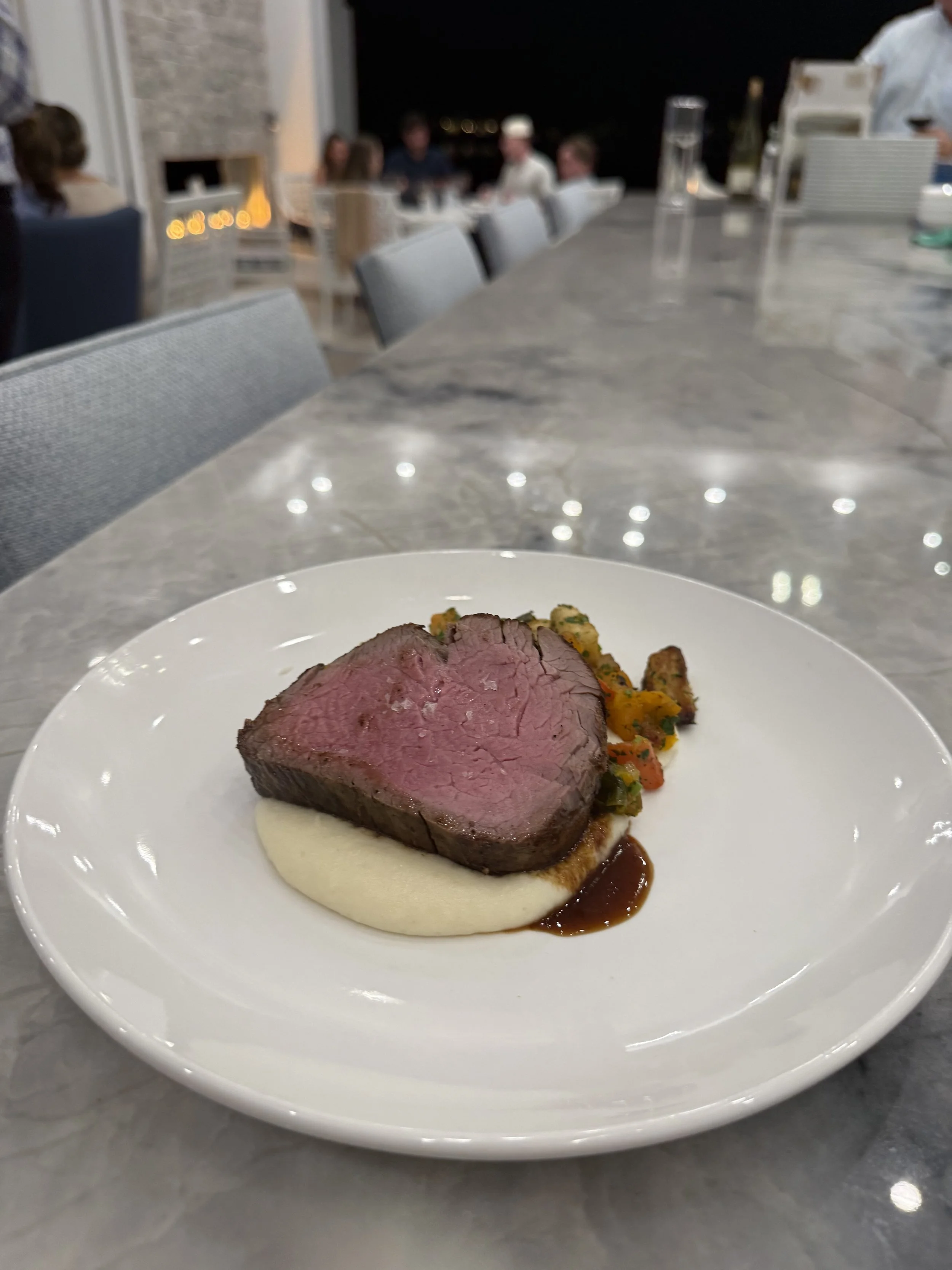 Plate with medium-rare steak, mashed potatoes, and colorful vegetables at a dinner table.