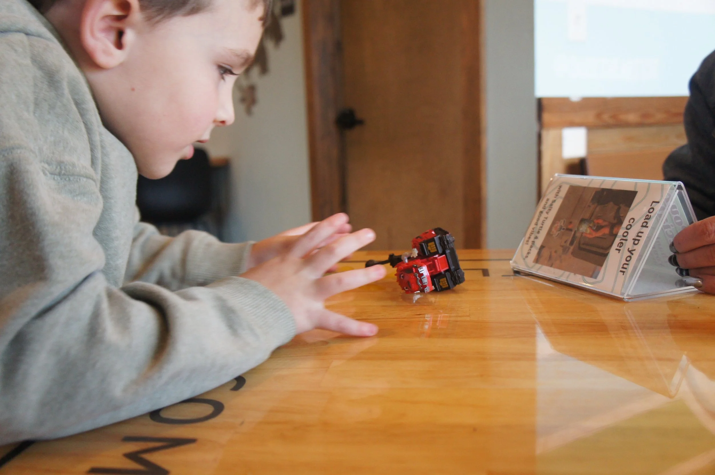 A young boy in a gray hoodie playing with a small red toy car on a wooden table, with a person holding a clear plastic display stand nearby.