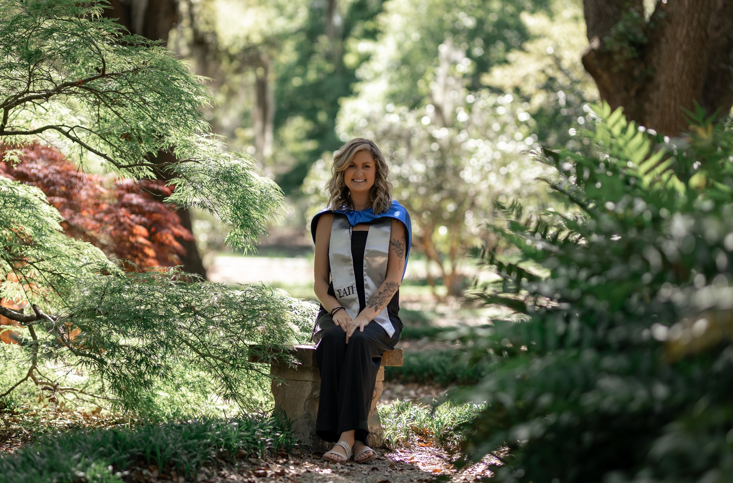 A young woman in a graduation cap and gown sitting on a stone bench in a lush, green park with trees and shrubs.