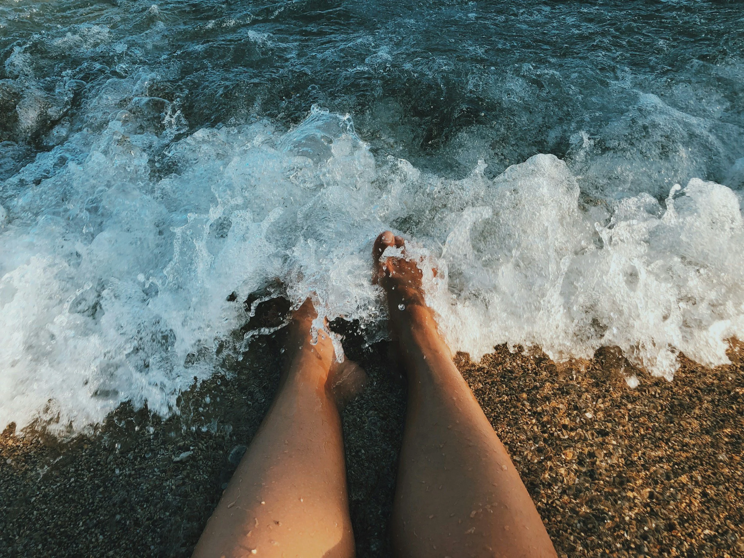 Person sitting on a rocky beach with their legs in the ocean, waves crashing around their feet.