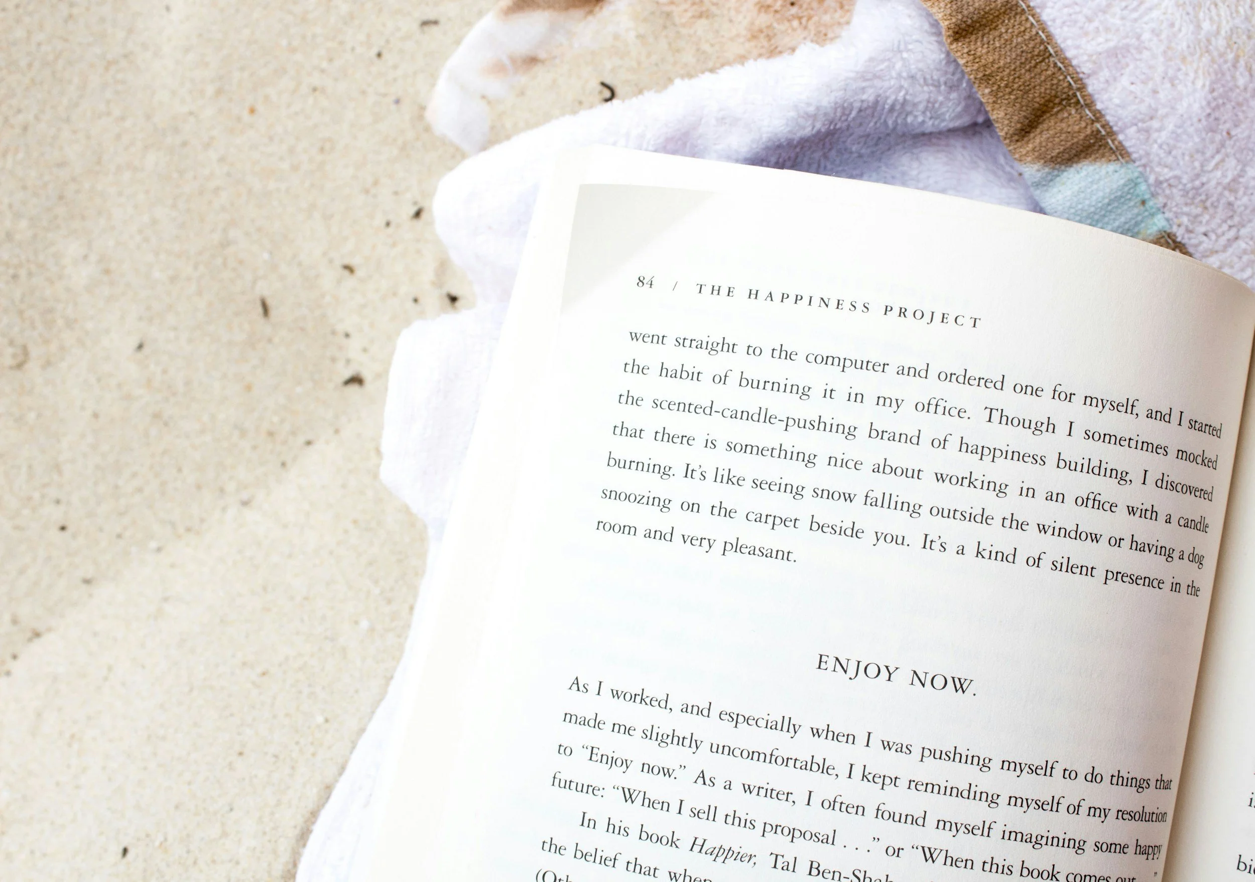 Open book on a white towel at the beach with sand visible nearby.