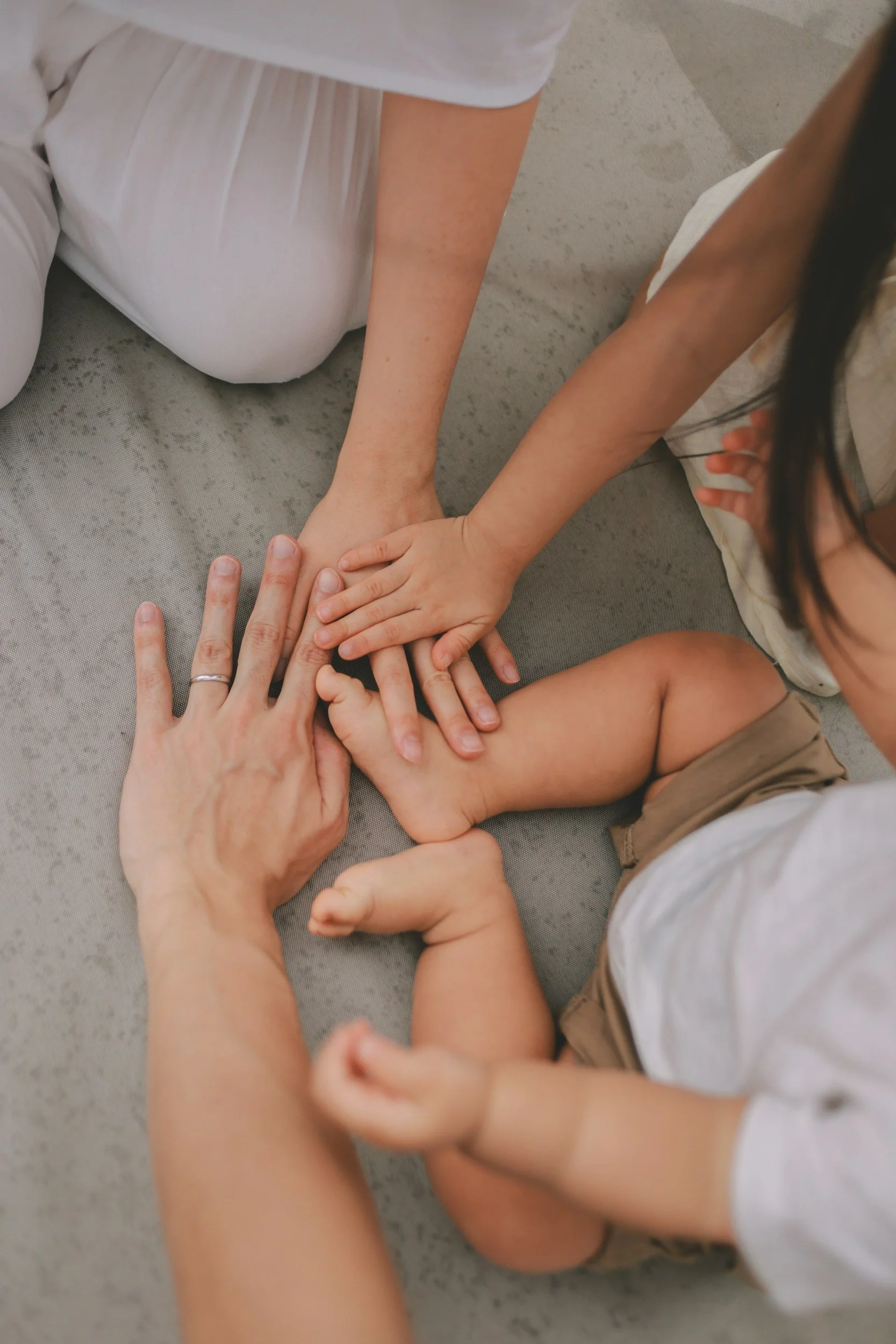 Three children and an adult are sitting on a gray carpet, stacking their hands together in a gesture of teamwork or friendship.