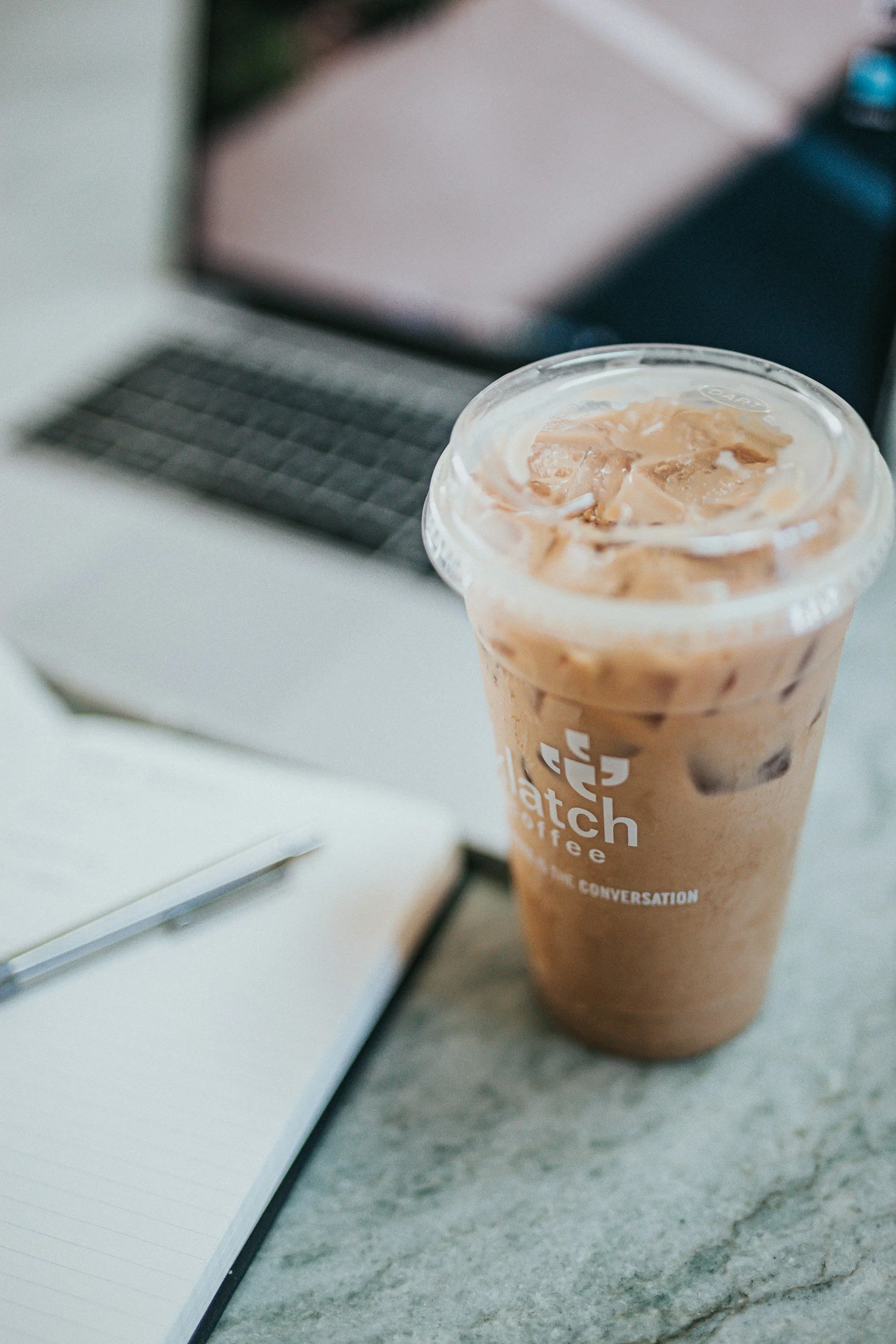 A tall plastic cup filled with iced coffee, placed on a marble surface, with a notebook and a laptop in the background.