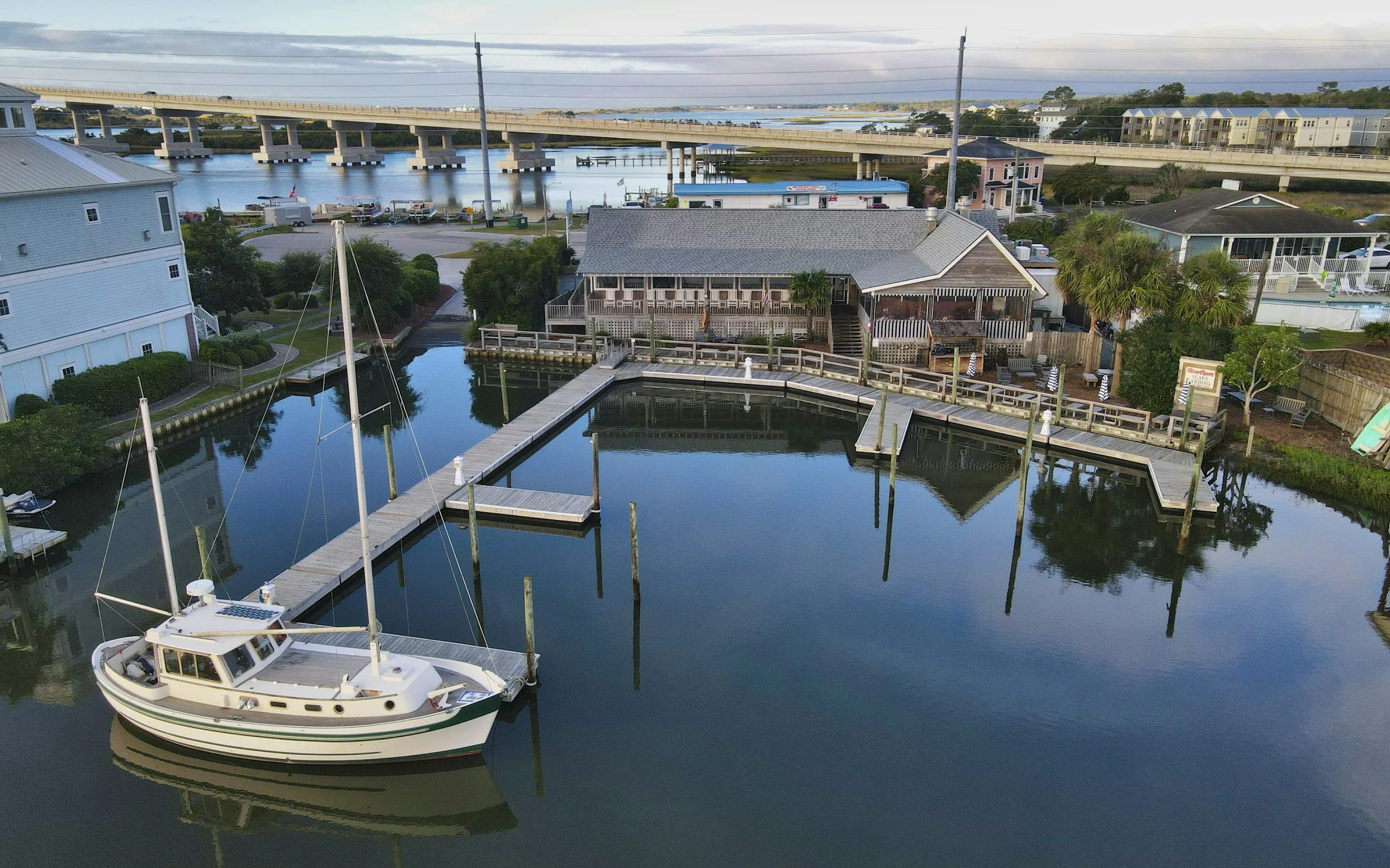 A marina with a white motorboat docked at a wooden pier, surrounded by calm water reflecting nearby buildings, trees, and a partly cloudy sky, with a bridge and waterway in the background