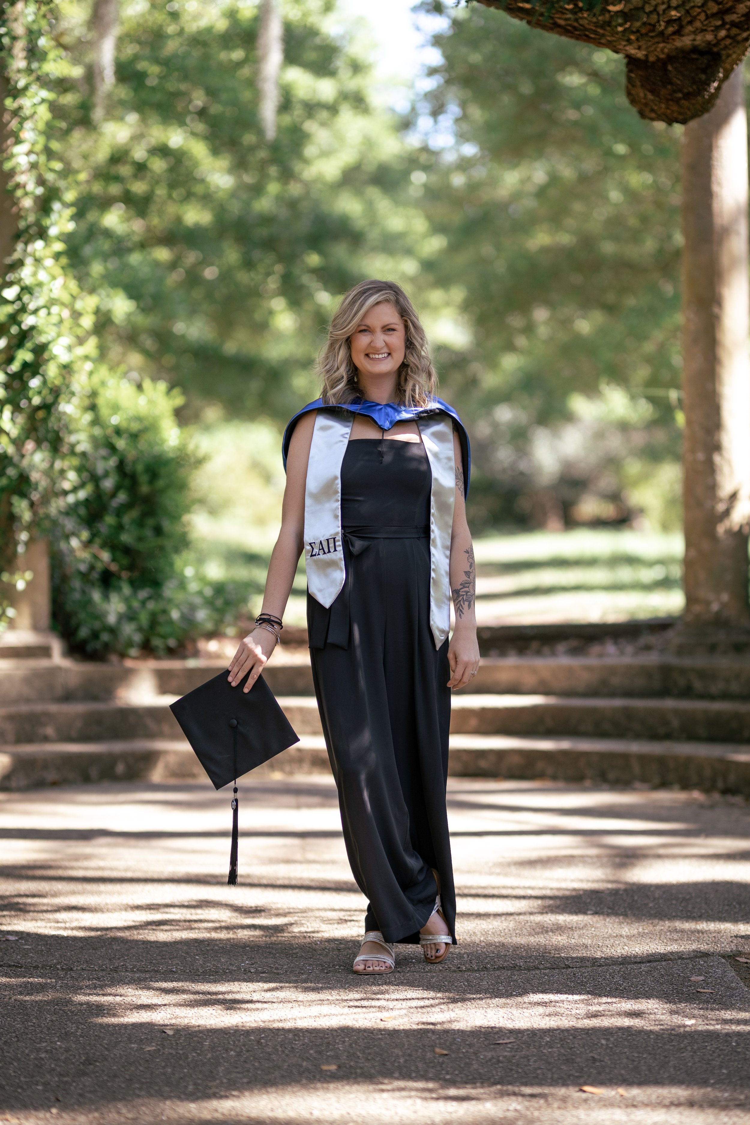 A woman in a graduation cap and gown standing on a pathway outdoors, smiling, with trees and greenery in the background.
