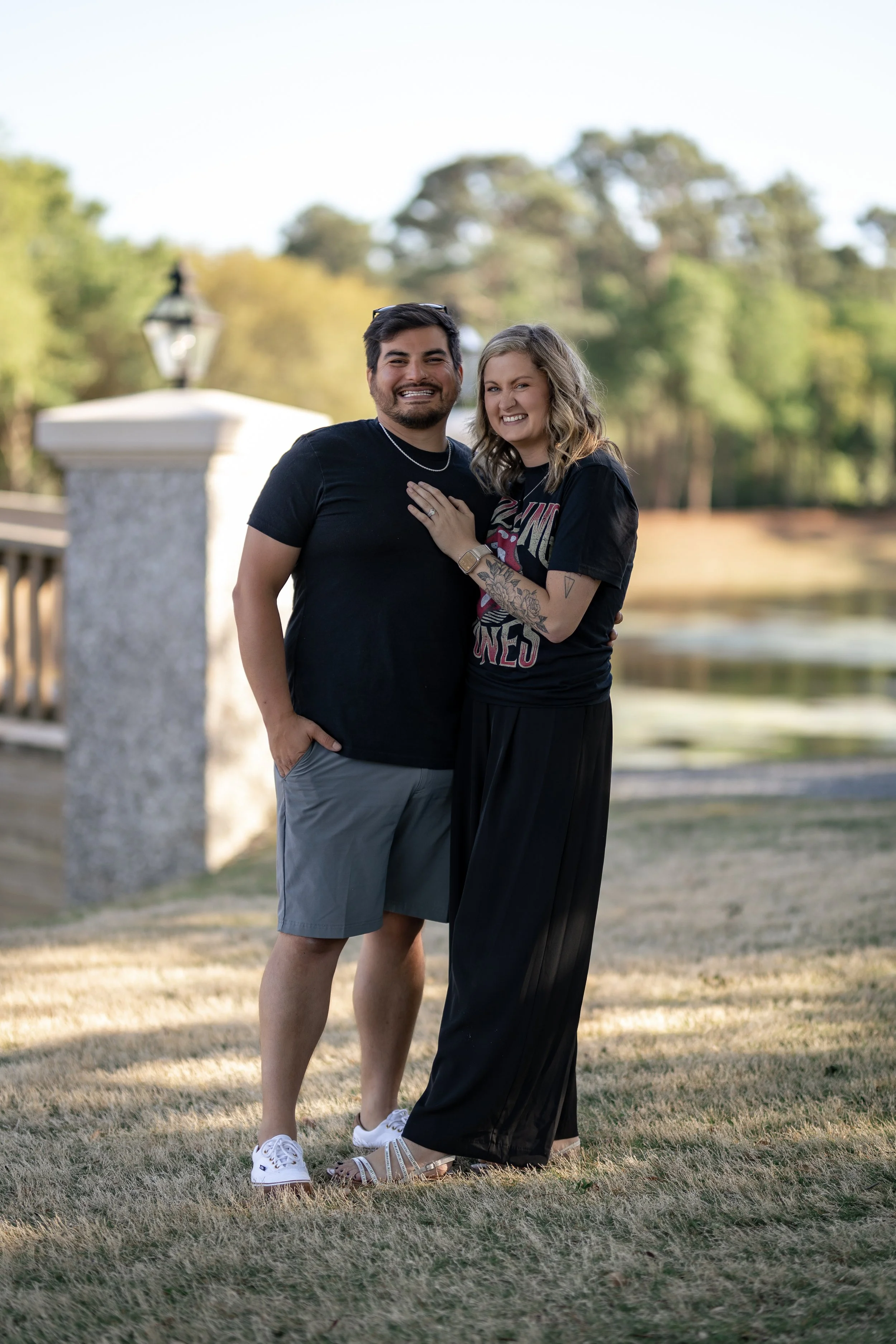 A happy couple standing outdoors on a grassy area near a bridge and pond, smiling at the camera.