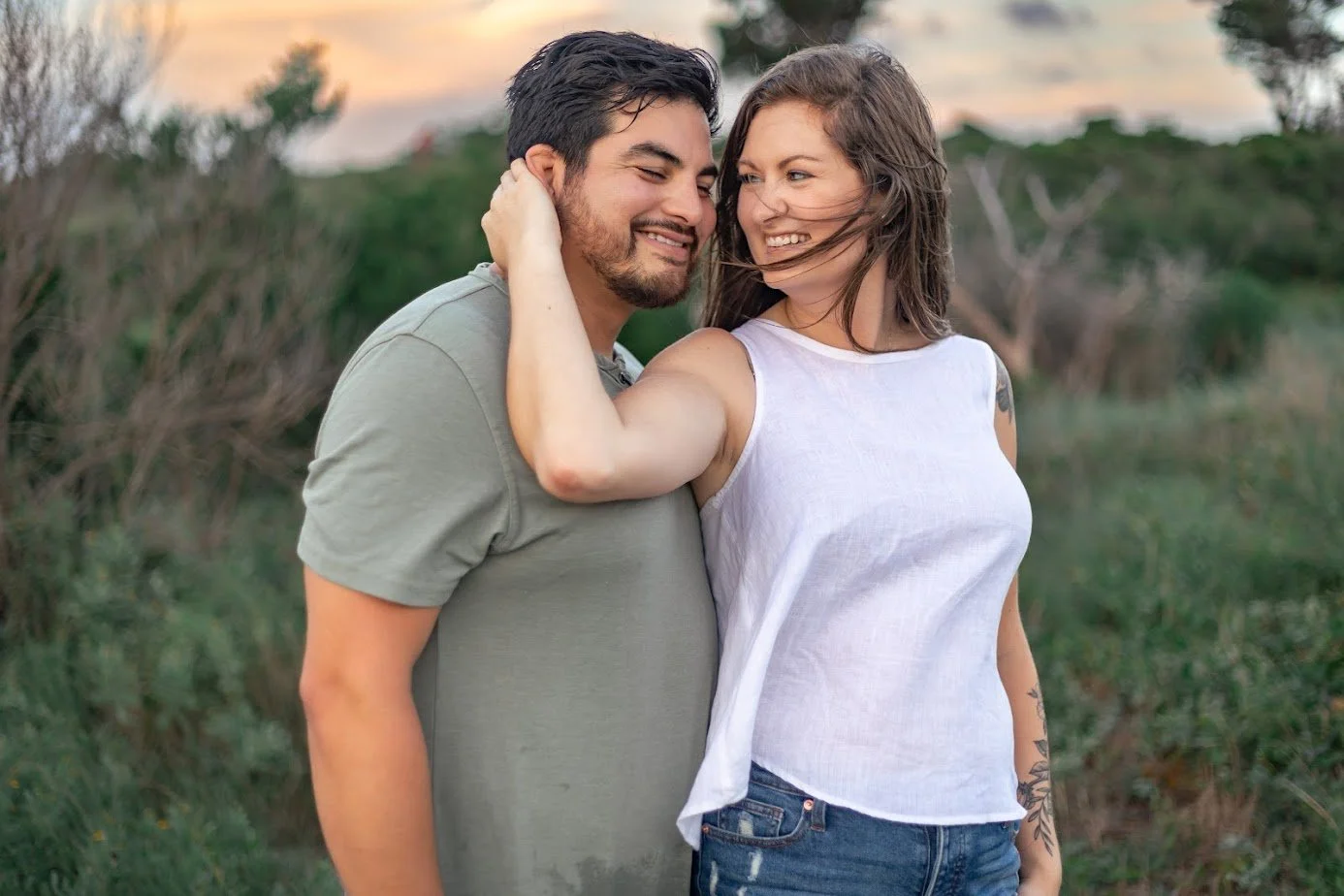 A happy couple stands close outdoors during sunset, smiling and embracing each other.