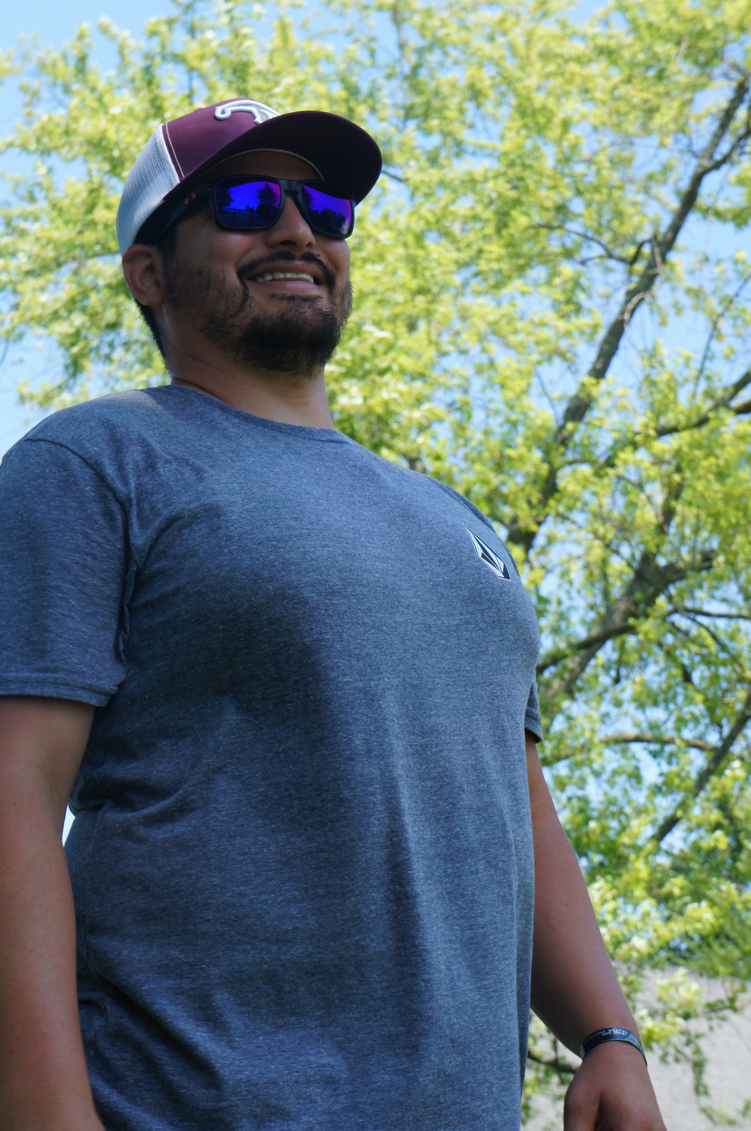A smiling man wearing sunglasses, a baseball cap, and a dark gray t-shirt, standing outdoors with green trees and a blue sky in the background.