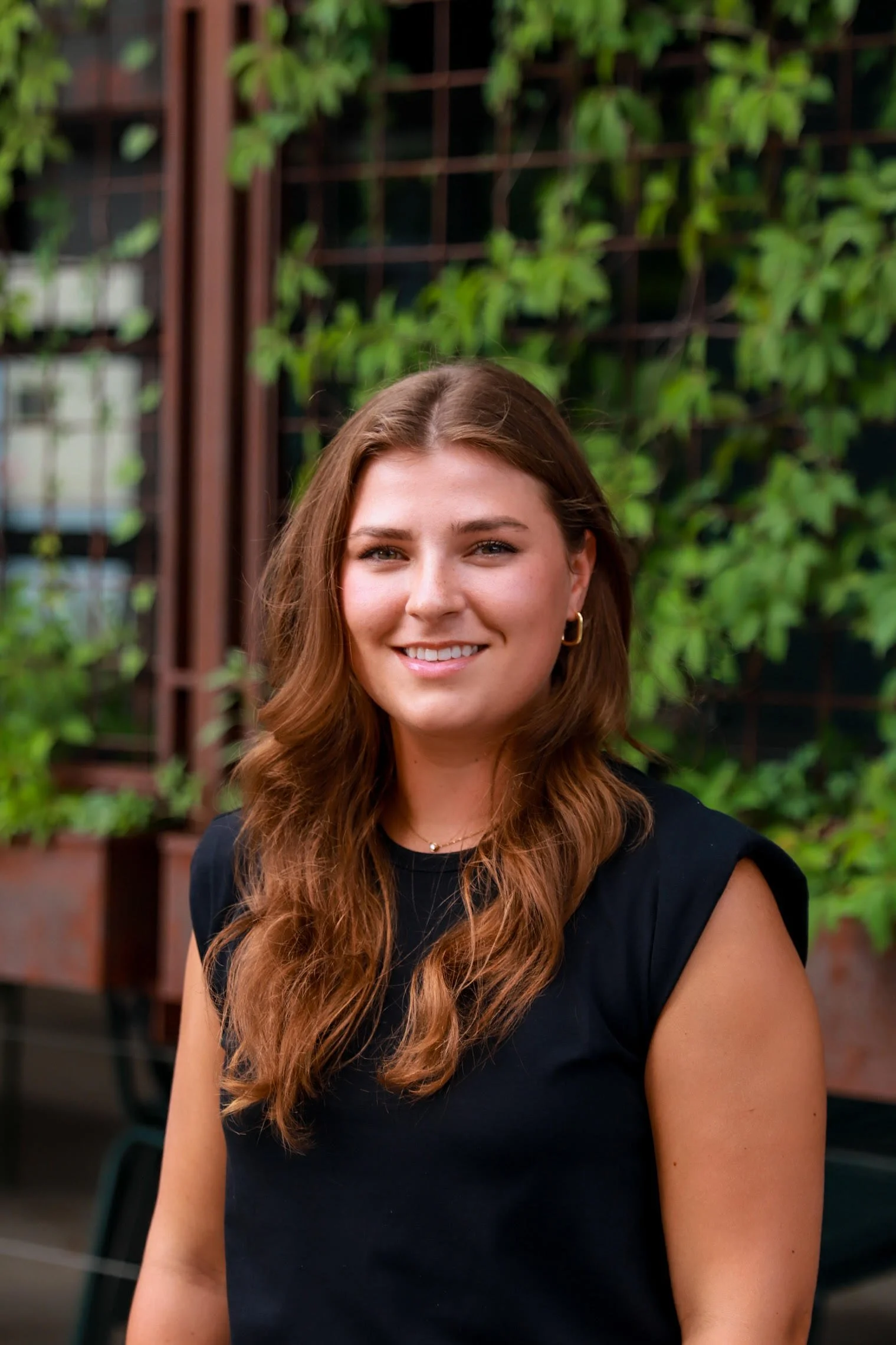 A young woman with long, wavy brown hair, wearing a black sleeveless top, smiling outdoors in front of a green leafy background.