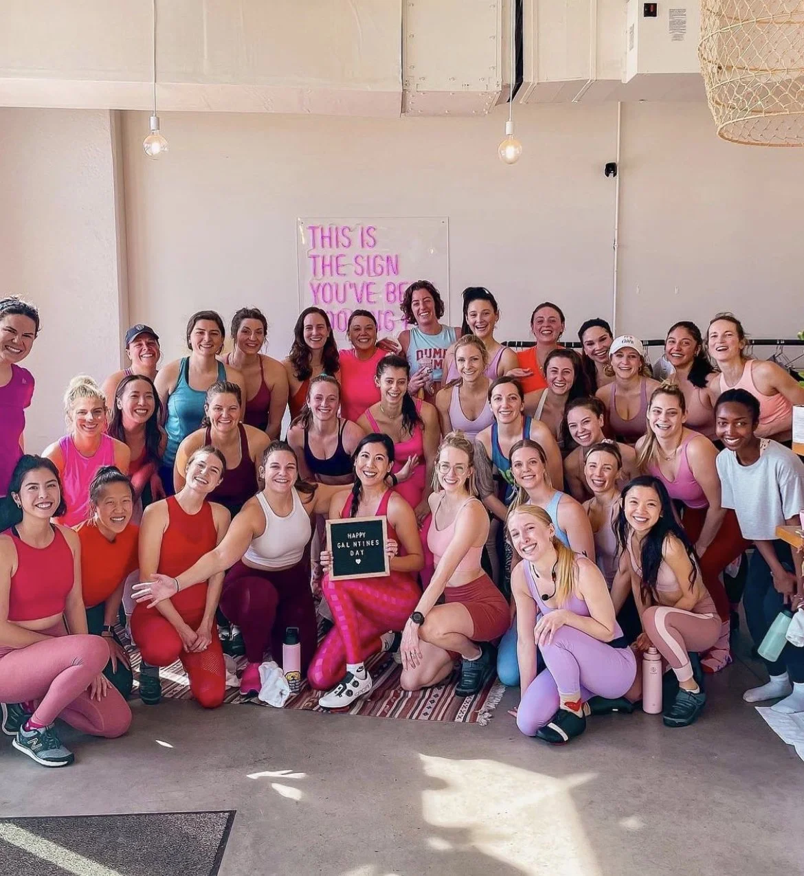 A large group of women dressed in colorful workout clothes, smiling and posing for a photo in a fitness or dance studio with a sign behind them that reads 'This is the sign you've been looking for.' One woman in the front holds a small sign that says 'Happy Galentines Day'.