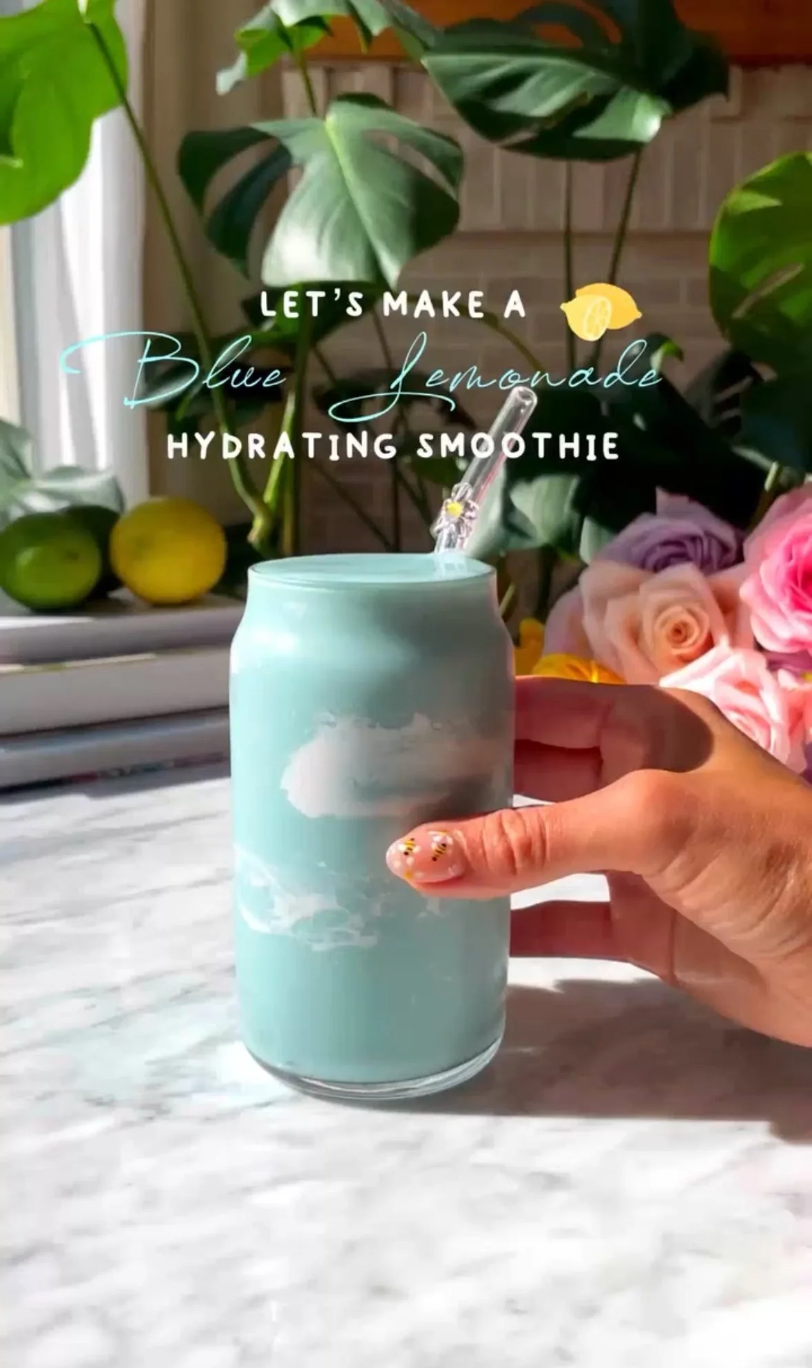 A hand holding a light blue can with a straw, surrounded by green plants, pink roses, and lemons, with a text overlay that reads "Let's make a blue lemonade hydrating smoothie."
