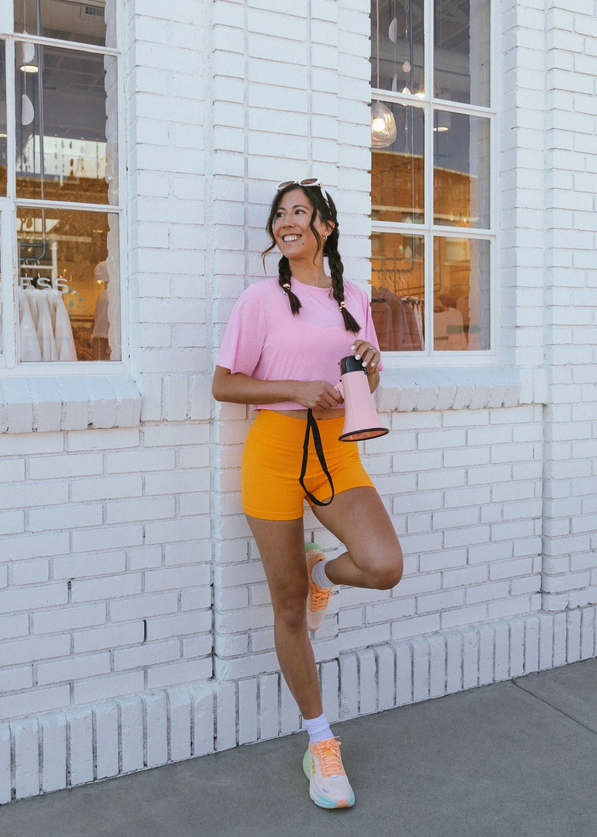 A woman wearing a pink t-shirt, yellow shorts, and colorful sneakers standing against a white brick wall, holding a pink megaphone, with sunglasses on her head, smiling and looking away.