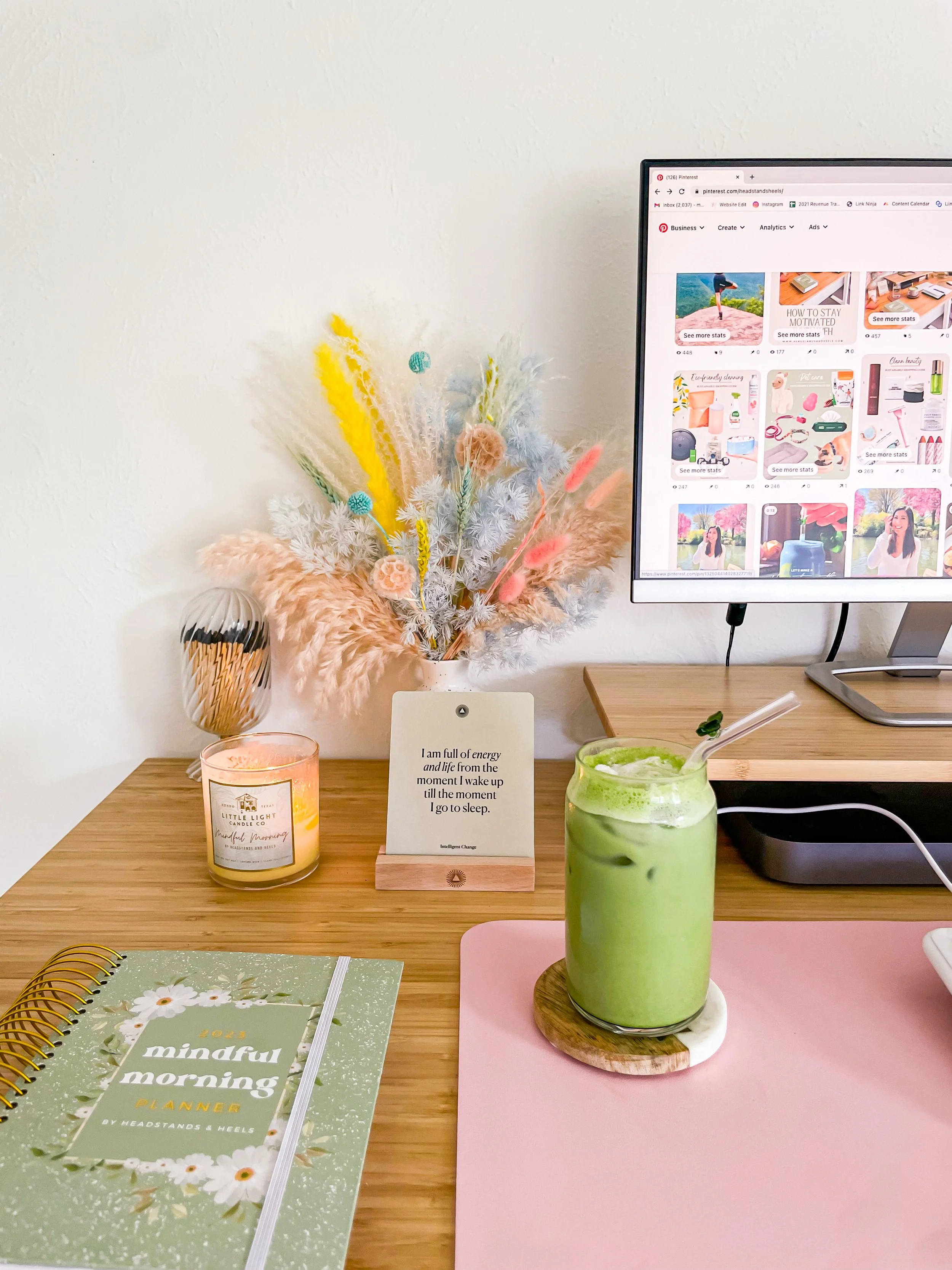 Desk setup with a pink drink on a coaster, a green smoothie with a straw, a planner, a lit candle, a decorative vase with pastel-colored dried flowers, a framed quote, and a computer monitor showing a Pinterest page.