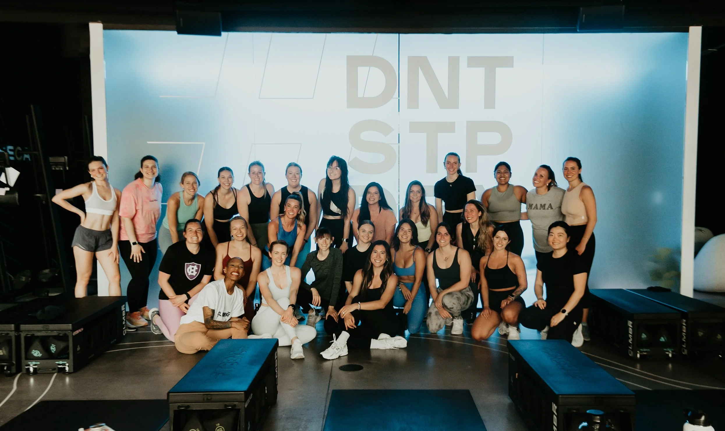 Group of women in workout attire smiling for a photo on a fitness stage with a large screen behind them displaying the text "DNT STOP".
