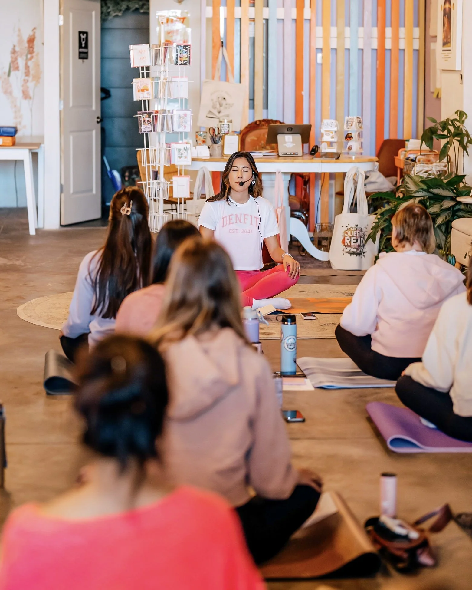 A woman giving a meditation or yoga class to a group of women sitting on yoga mats in a cozy, well-lit room with wooden floors and colorful walls.