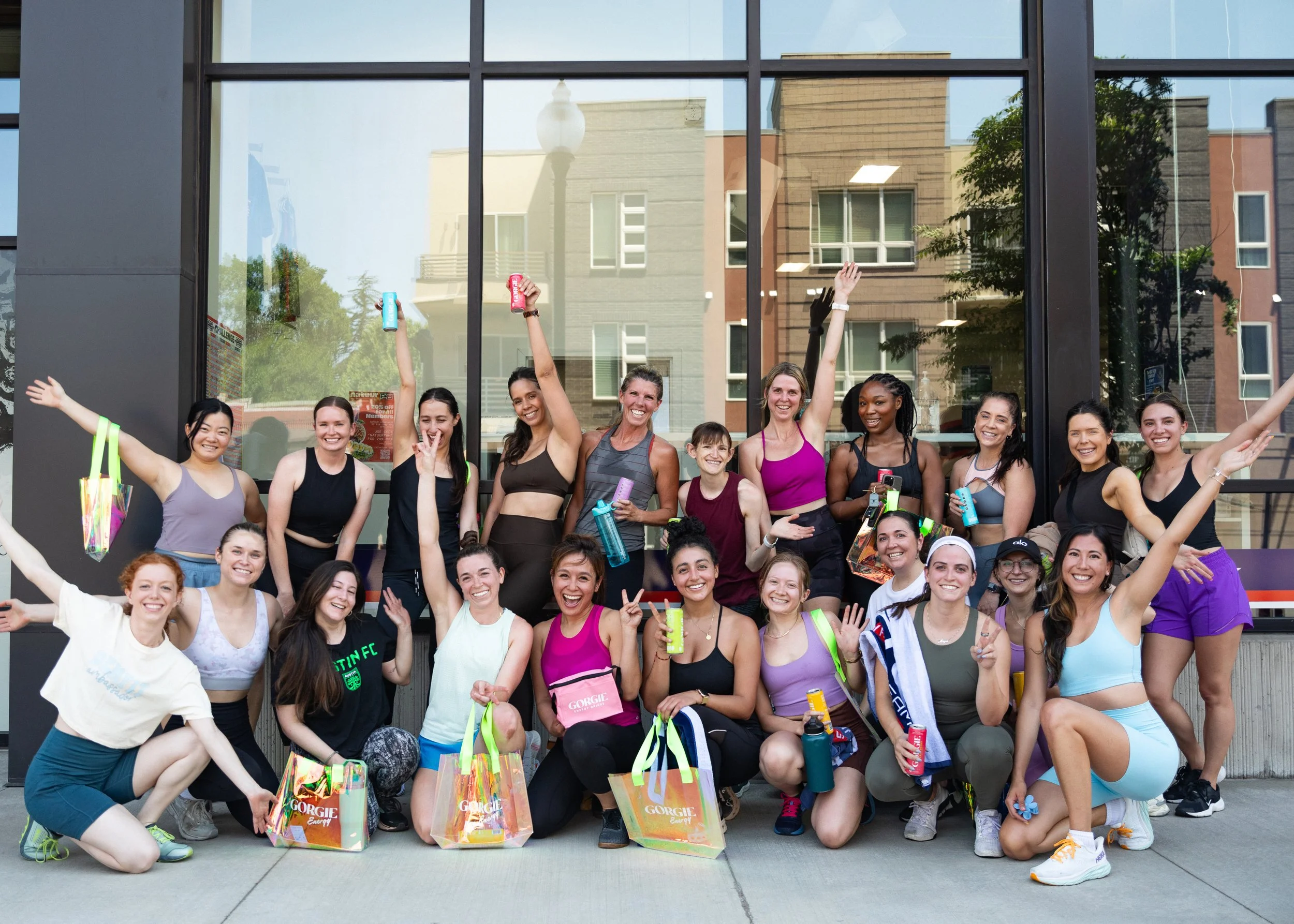 Group of women in athletic clothing celebrating after a workout outdoors, some holding water bottles and energy drinks, in front of a reflective building window.