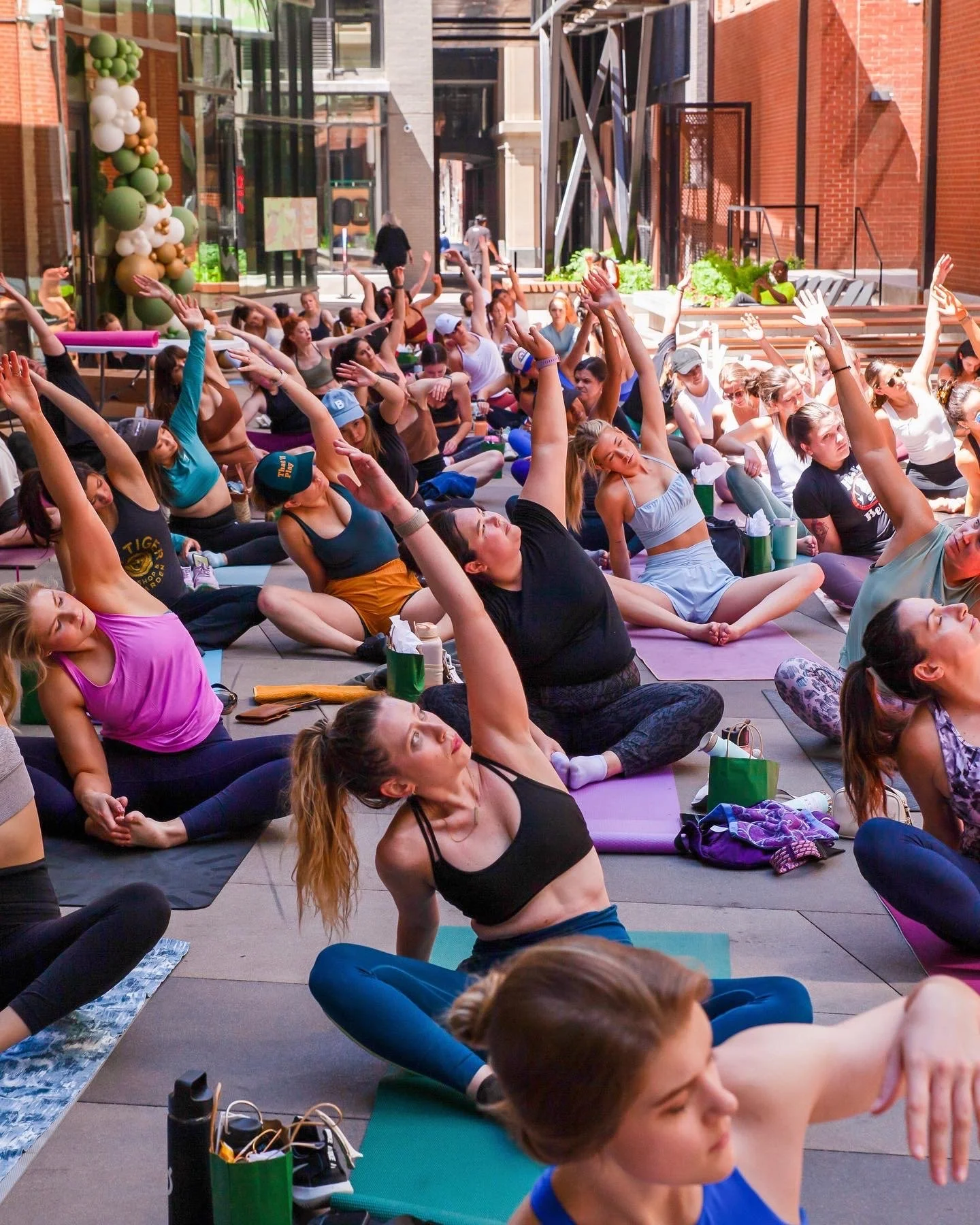 A large group of people participating in a yoga class outdoors in an urban area, sitting on yoga mats, stretching and practicing poses.